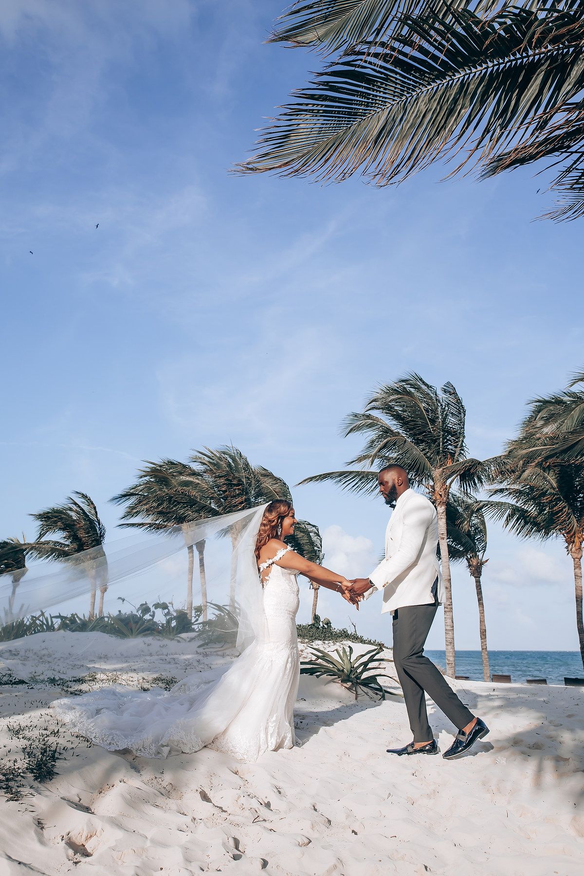 Wind creates dramatic movement with a wedding dress on a sandy beach with palm trees.