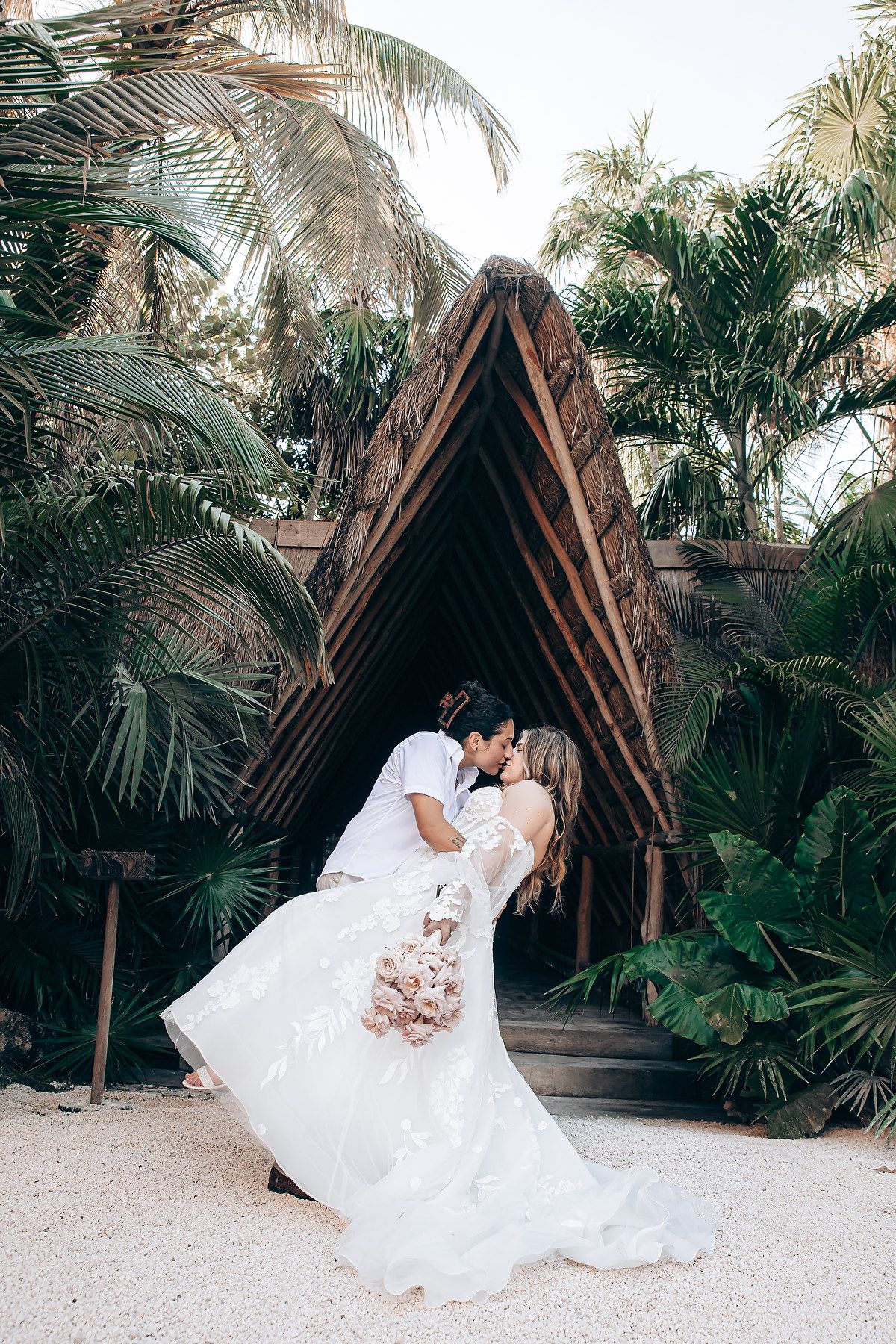 A romantic wedding couple poses in a tropical garden setting with palm trees and wooden structures.