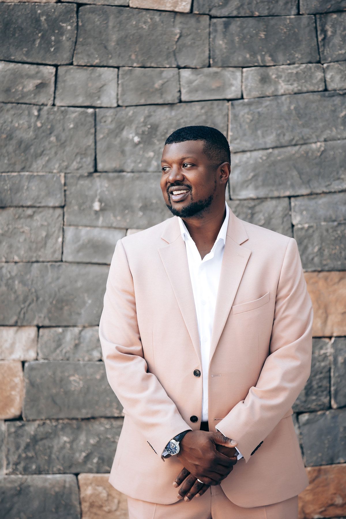 Person wearing light pink blazer poses against stone wall in formal portrait setting.