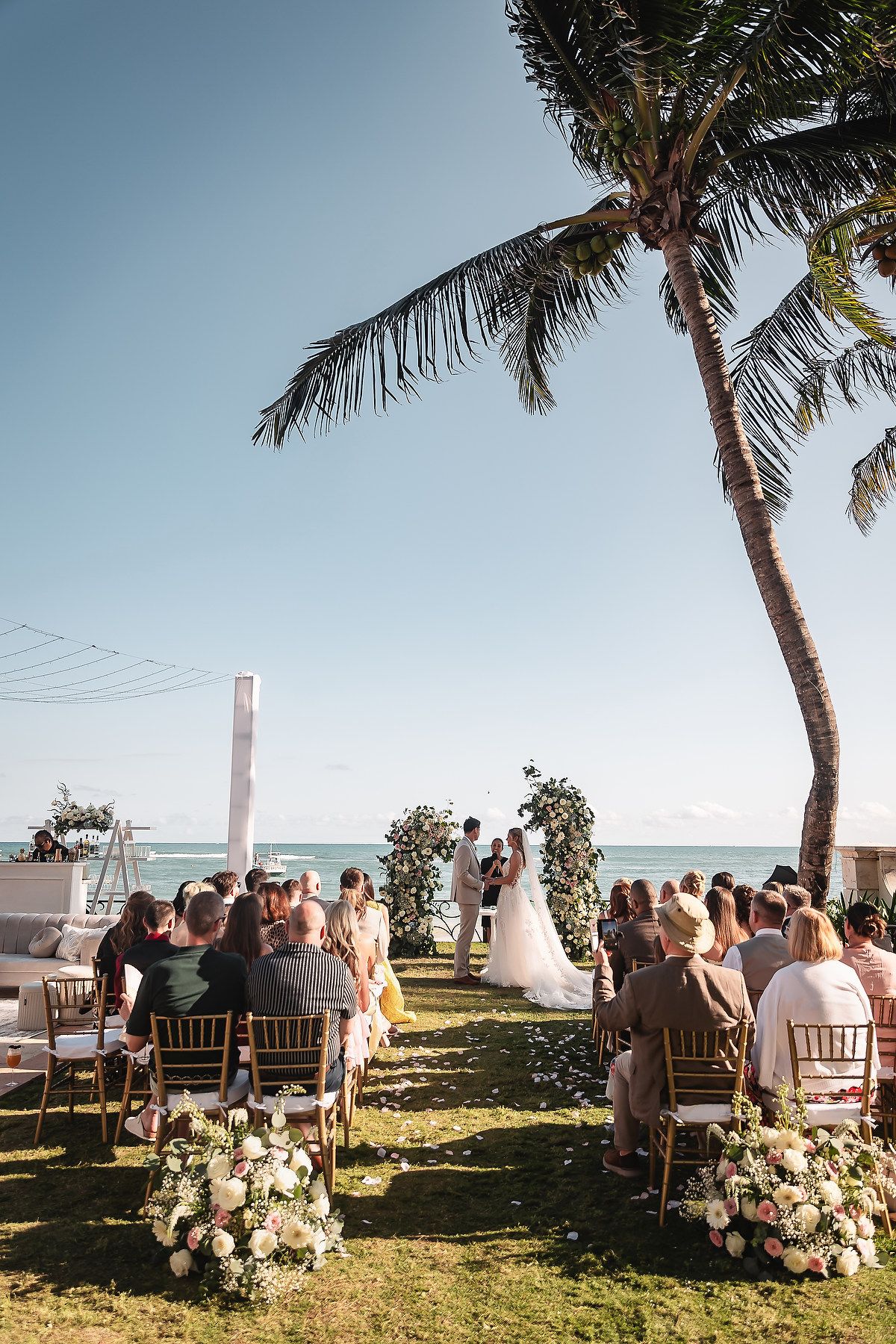 Beachside wedding ceremony setup with white chairs, floral arrangements, and palm trees against ocean backdrop.