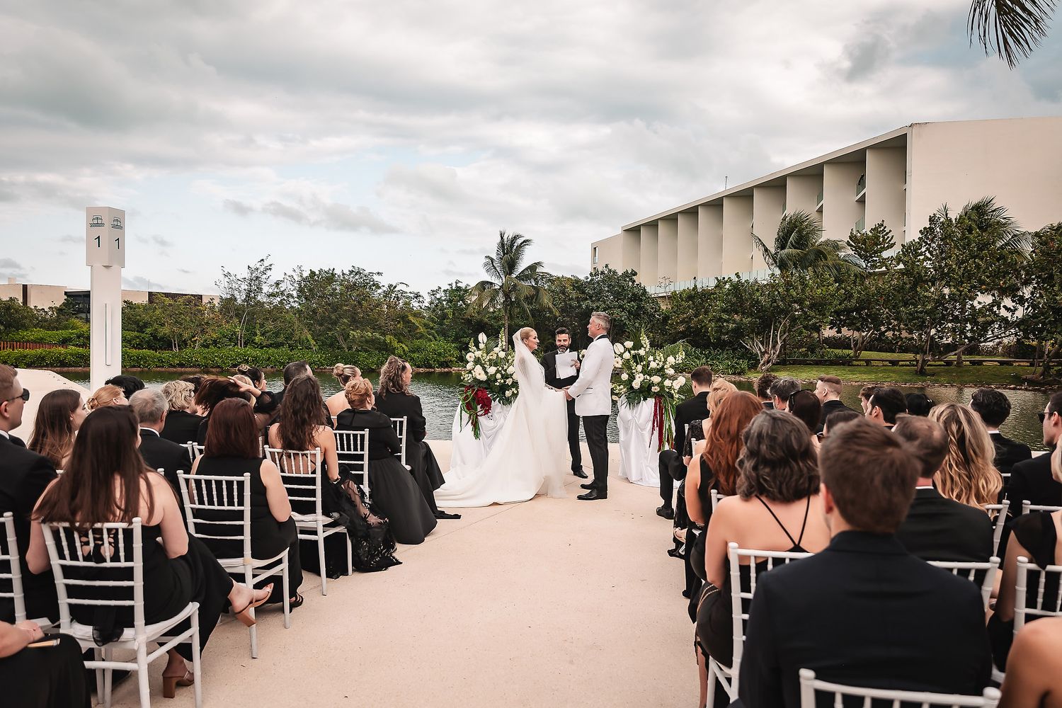 Large wedding party poses outdoors with palm trees and modern architecture in the background.