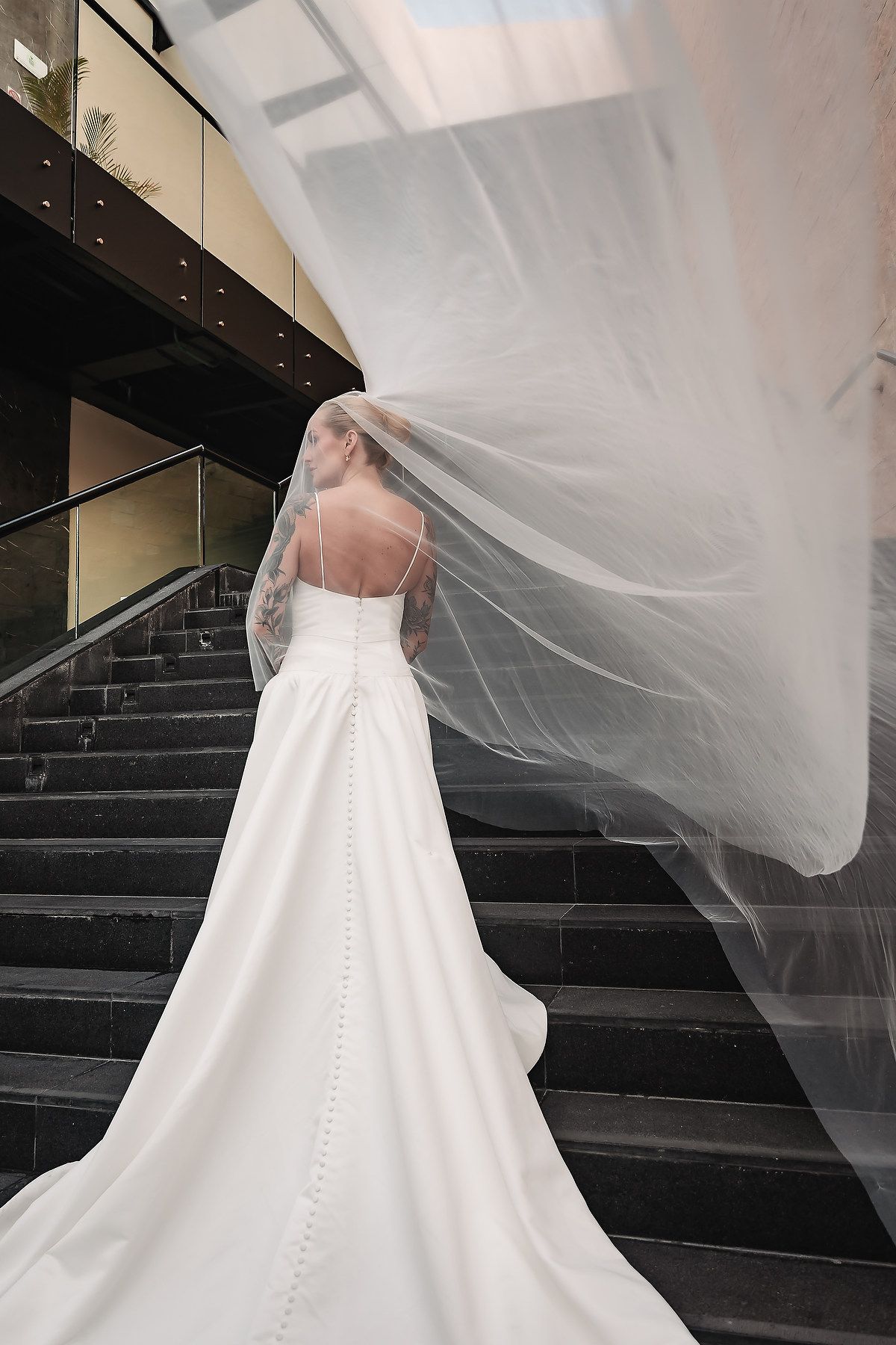 Elegant white wedding dress with flowing train and veil cascades down dark stairs.