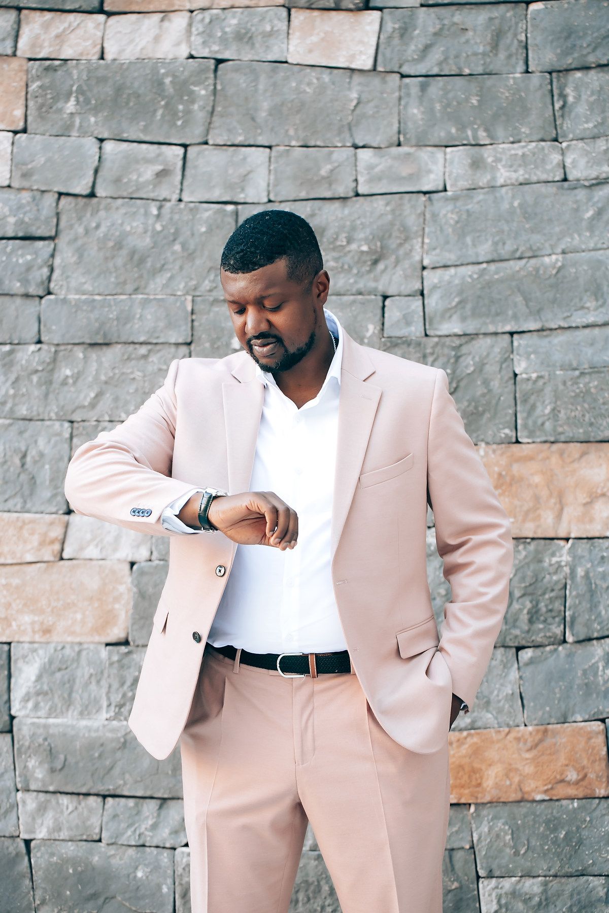 Person in light pink suit poses confidently against a modern stone wall background.
