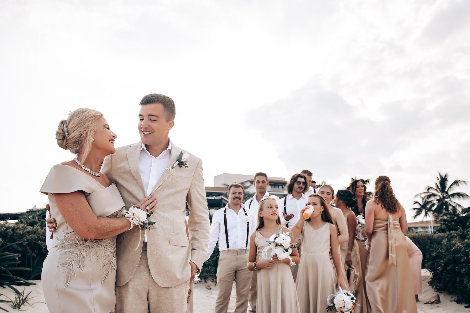 Wedding party in beige and white attire poses together on a sunny beach.