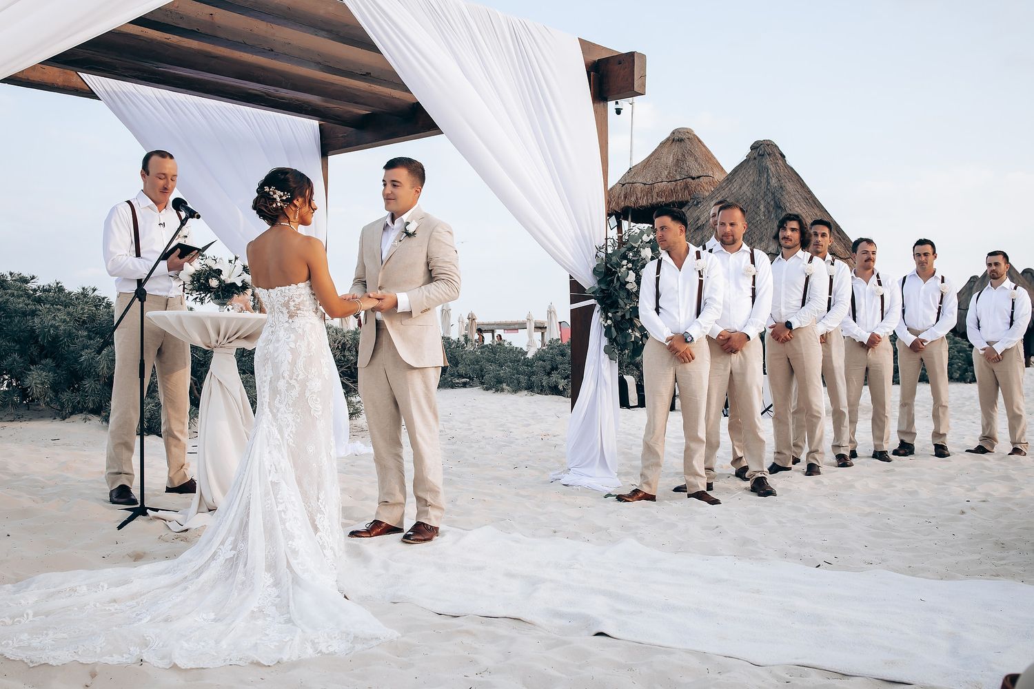 Wedding party poses on a sandy beach with palm trees and resort buildings in the background.