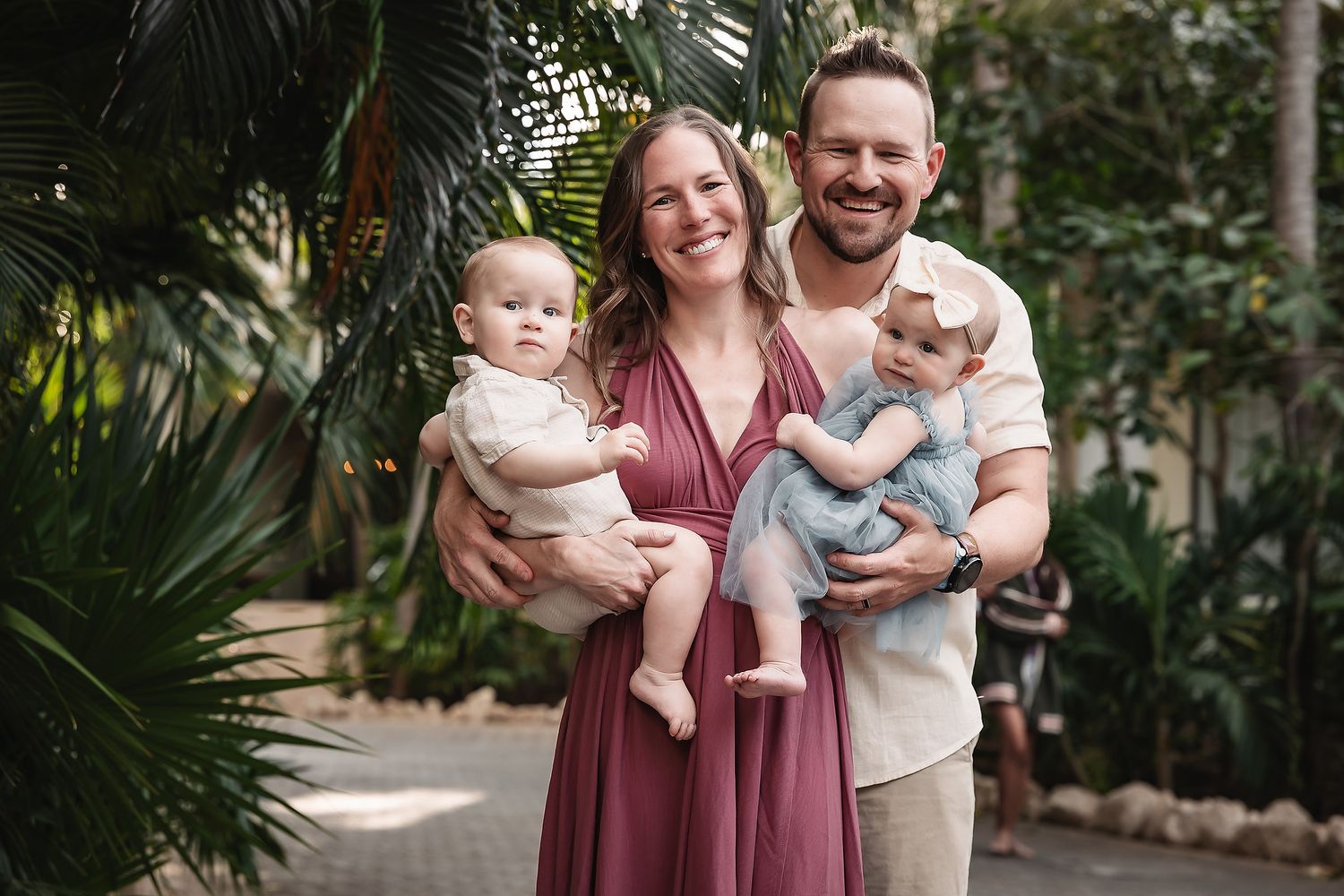 Happy family with two babies posing outdoors among lush tropical greenery, smiling couple holding infants in natural light.