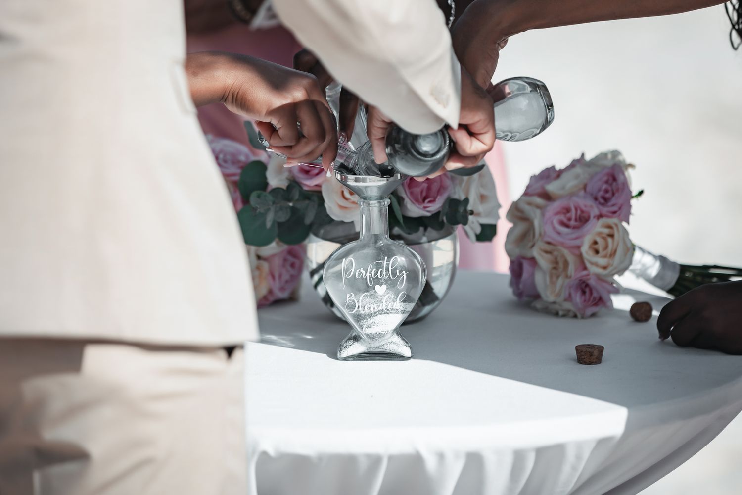Couple pouring sand into engraved unity vase during wedding ceremony with pink floral bouquet nearby.