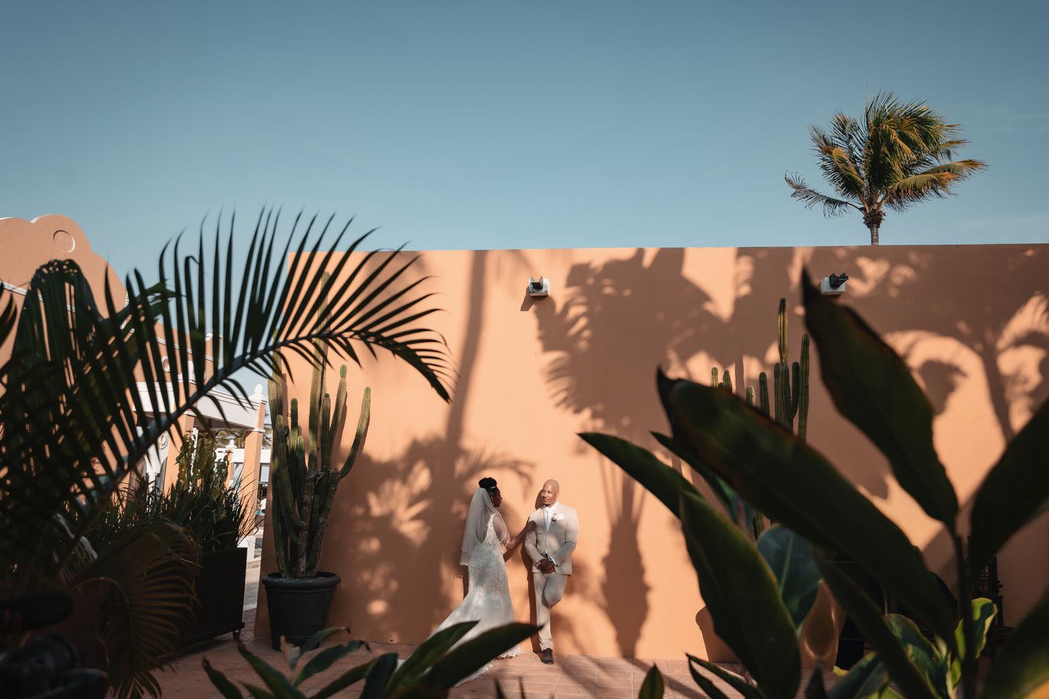 Two people in white robes walk beside a terracotta wall with palm tree shadows in a sunny tropical setting.