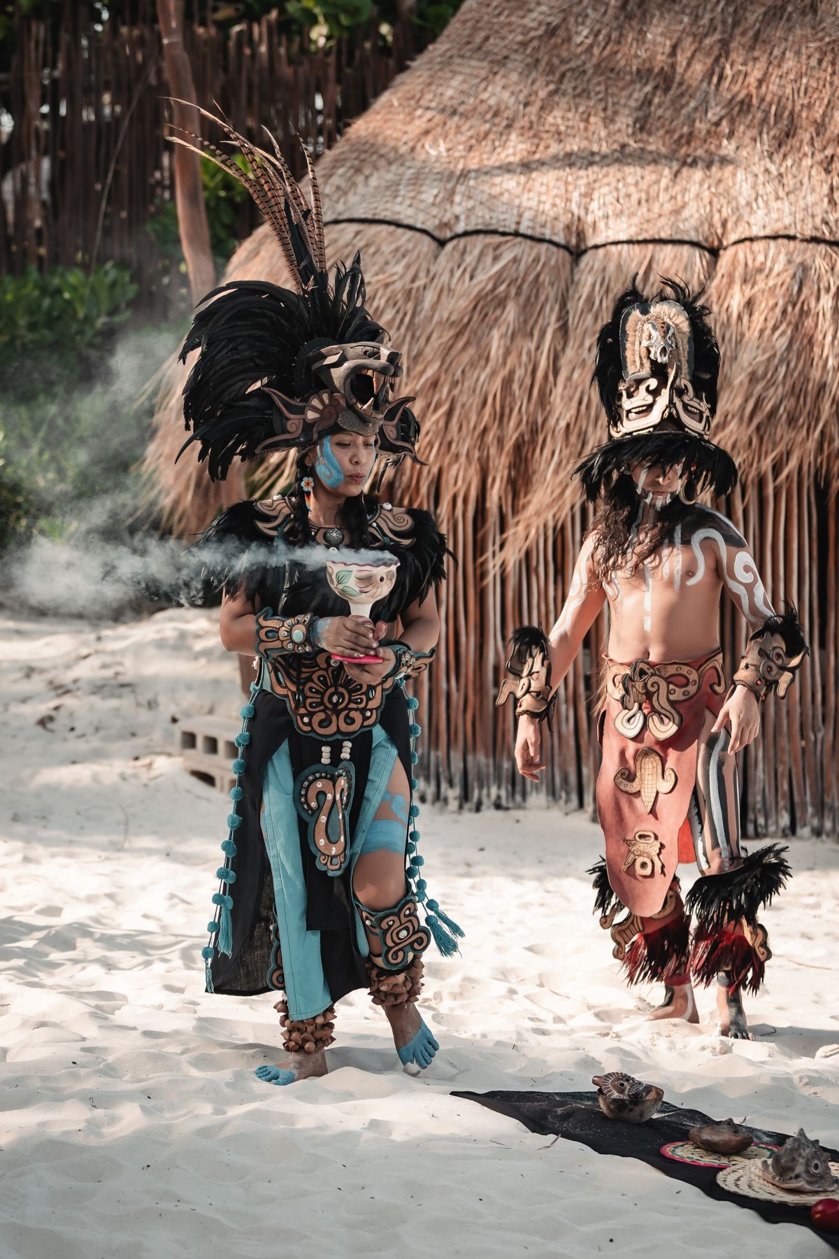 Two performers in elaborate Mayan costumes with feathered headdresses during a traditional ceremony on a sandy beach.