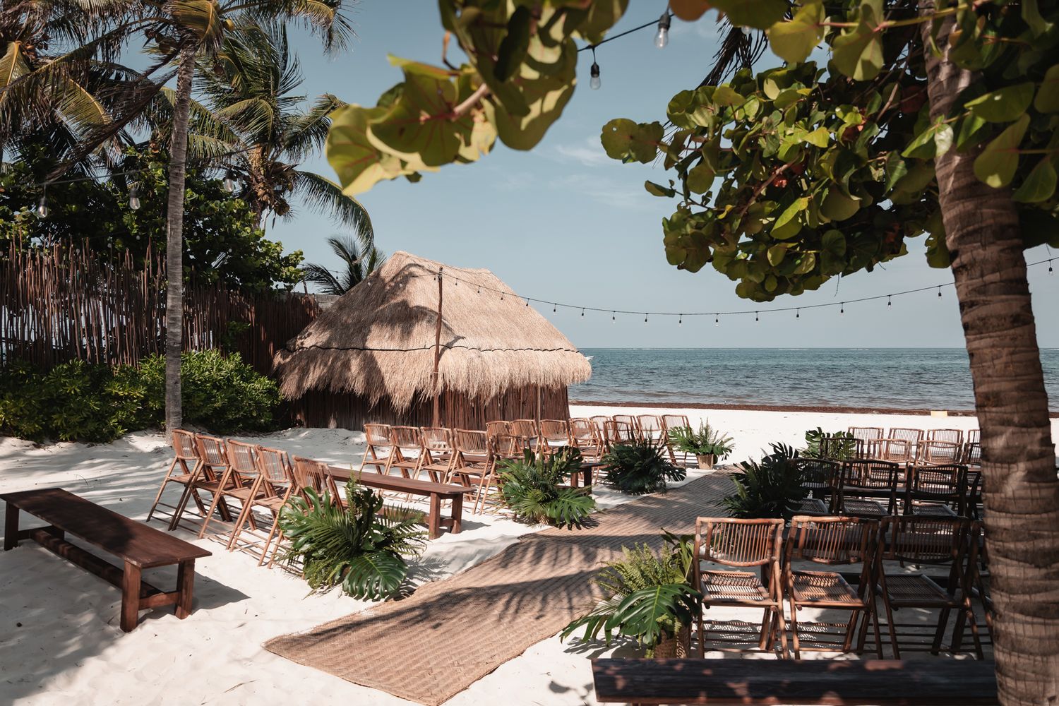 Beachfront wedding ceremony setup with wooden chairs, tropical plants, and a thatched palapa on a white sand beach.