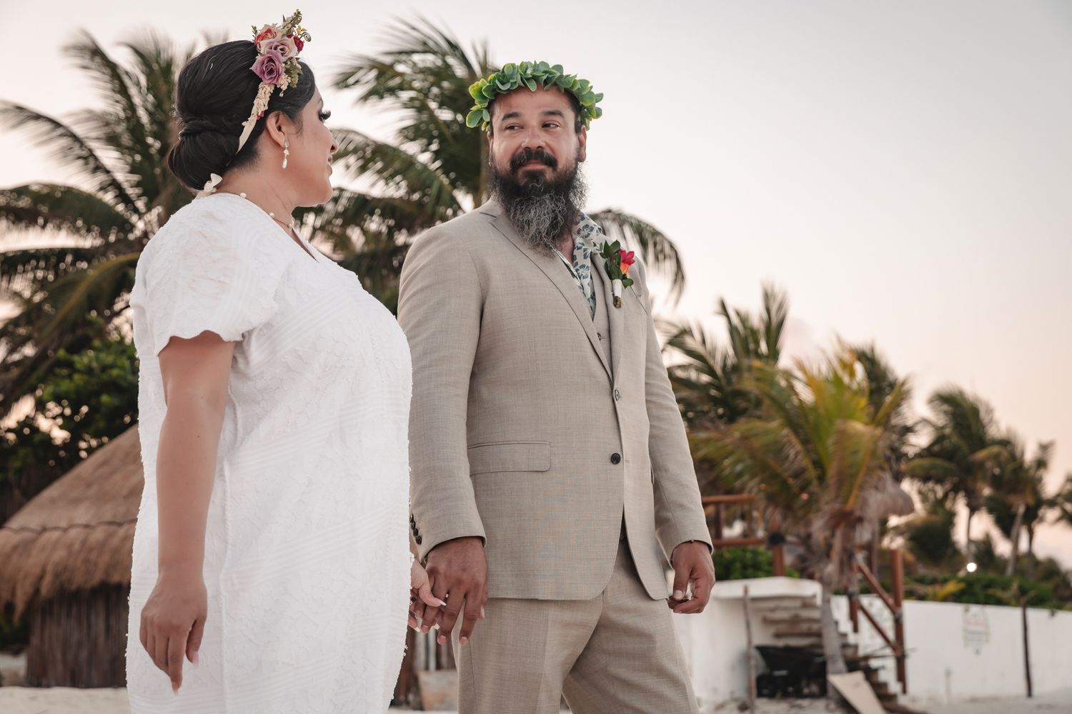 Couple in floral crowns holding hands on a tropical beach, bride in white dress and groom in tan suit.