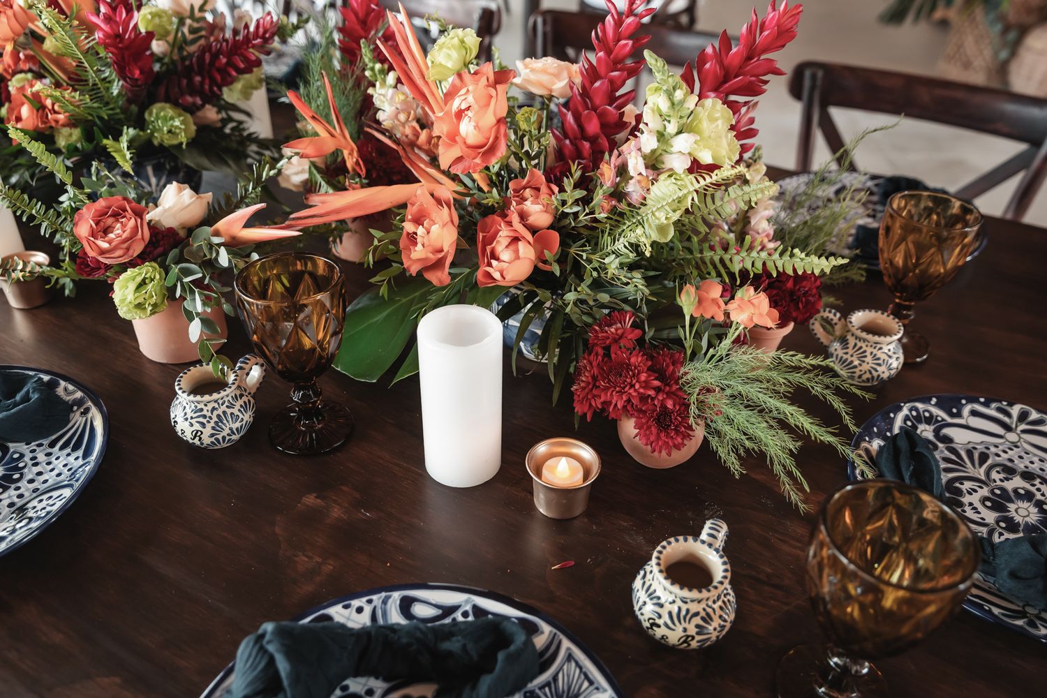 Elegant wedding tablescape with tropical floral centerpieces, candles, and dark blue patterned napkins on a wooden table.