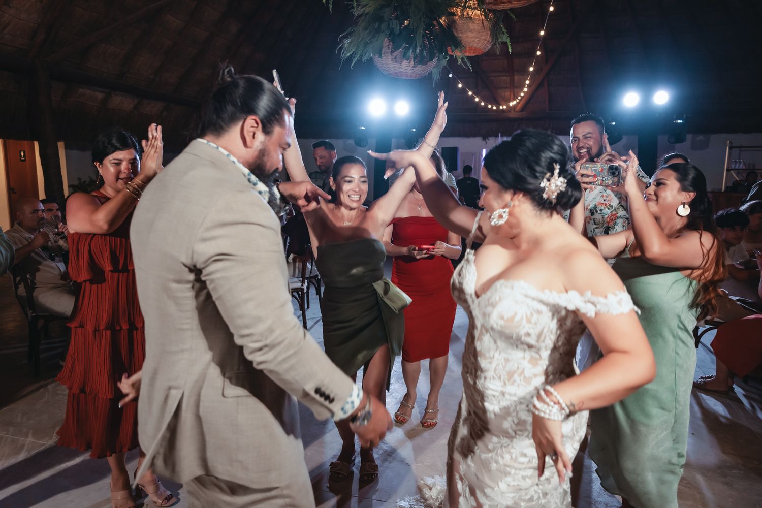 Bride and groom dancing joyfully with guests at a wedding reception, surrounded by cheering bridesmaids in colorful dresses.