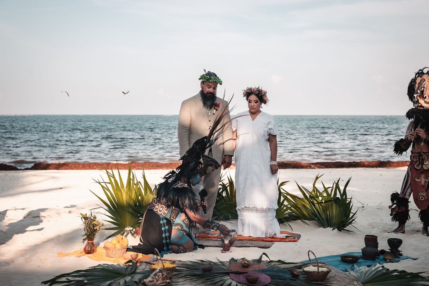 Couple in wedding attire on a beach with ocean backdrop, surrounded by cultural decorations and a costumed officiant.