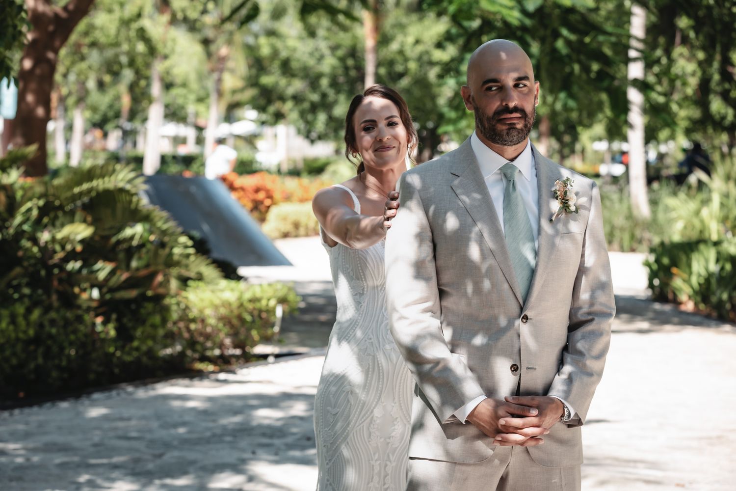Bride in white gown and groom in gray suit pose outdoors in a sunny park setting on their wedding day.