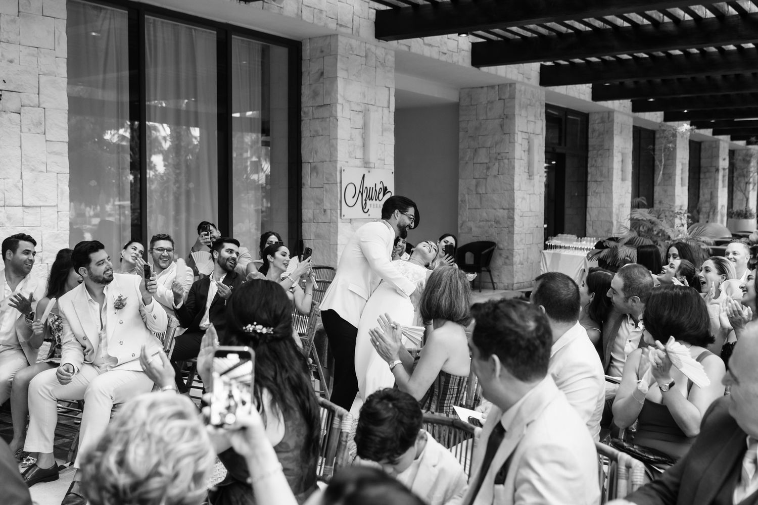 Black and white wedding photo of couple sharing a kiss surrounded by clapping guests at outdoor ceremony.
