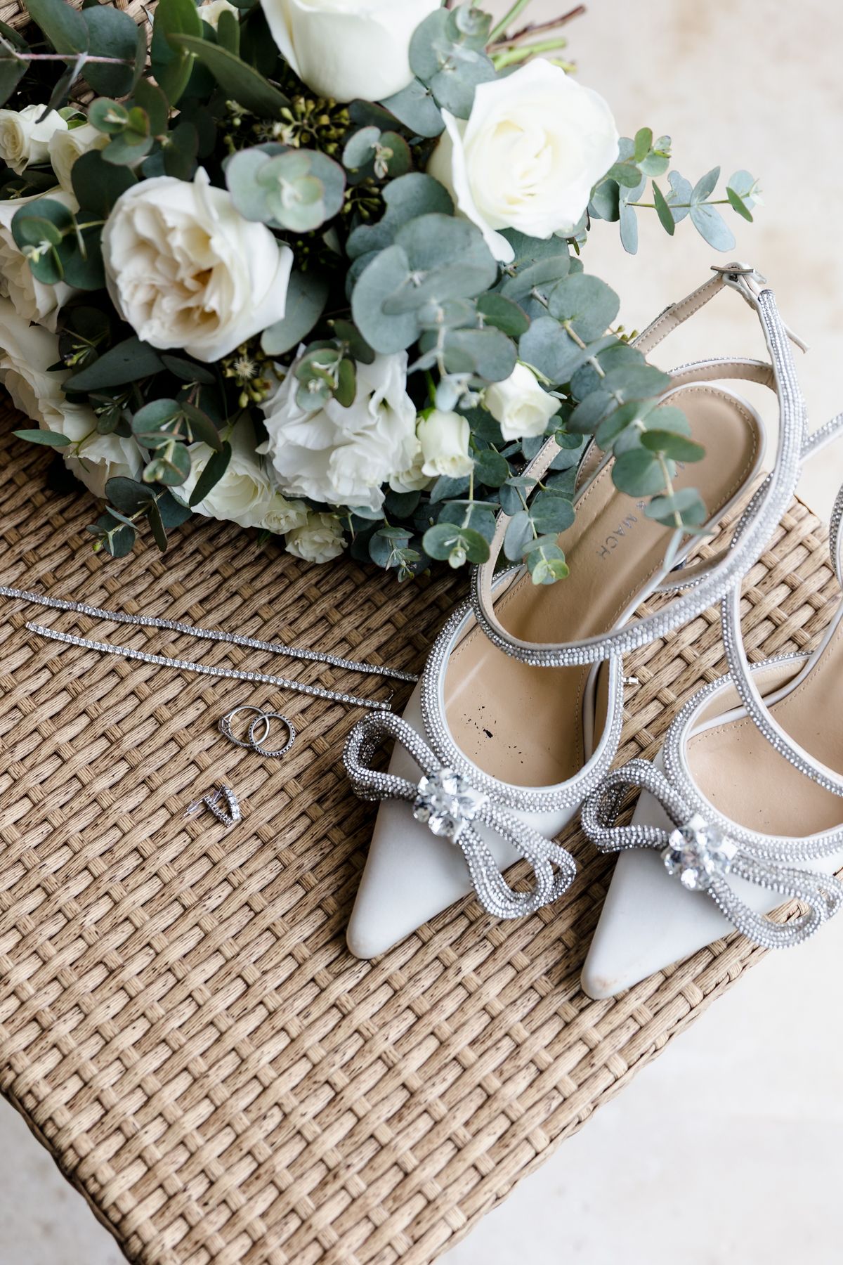 Wedding flat lay with crystal bow heels, white rose bouquet, rings, and necklace on wicker.