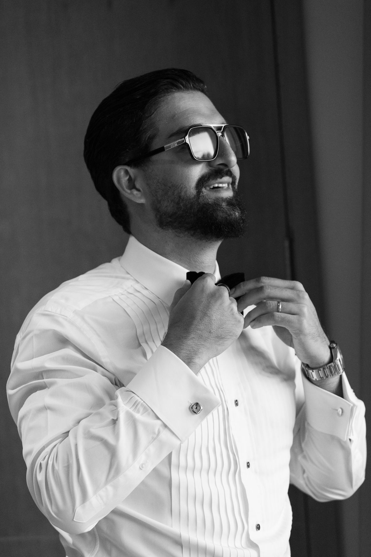 Bearded groom in white tuxedo shirt adjusting bow tie wearing glasses, black and white wedding photo.