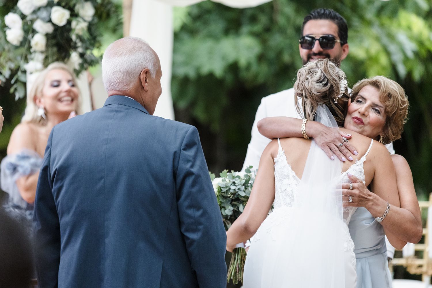 Bride receives warm hug from mother-in-law as groom watches during outdoor wedding ceremony.