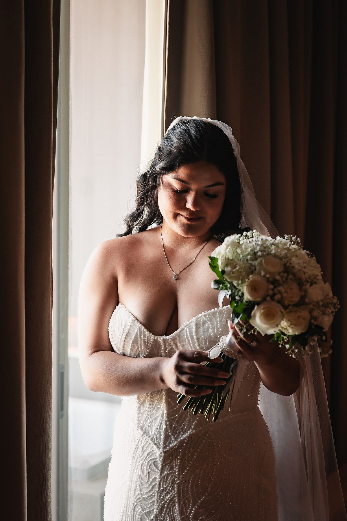 Elegant bride in lace gown with veil smiling at white rose bouquet by hotel window.
