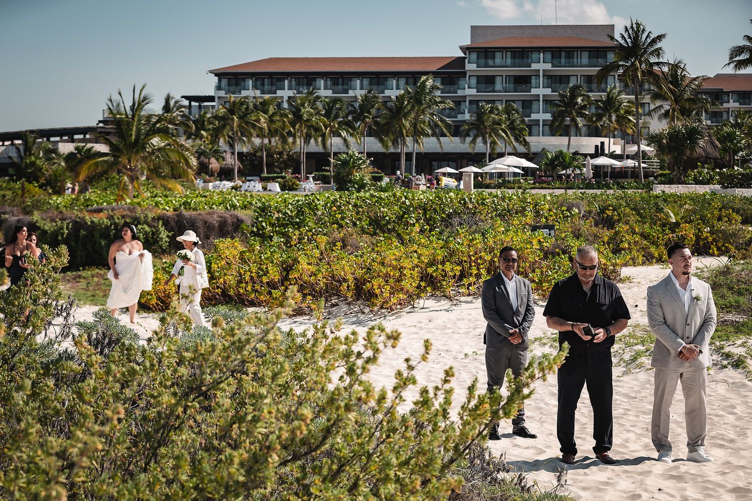 Beach wedding ceremony with guests and officiant on white sand, tropical resort and palm trees in background.