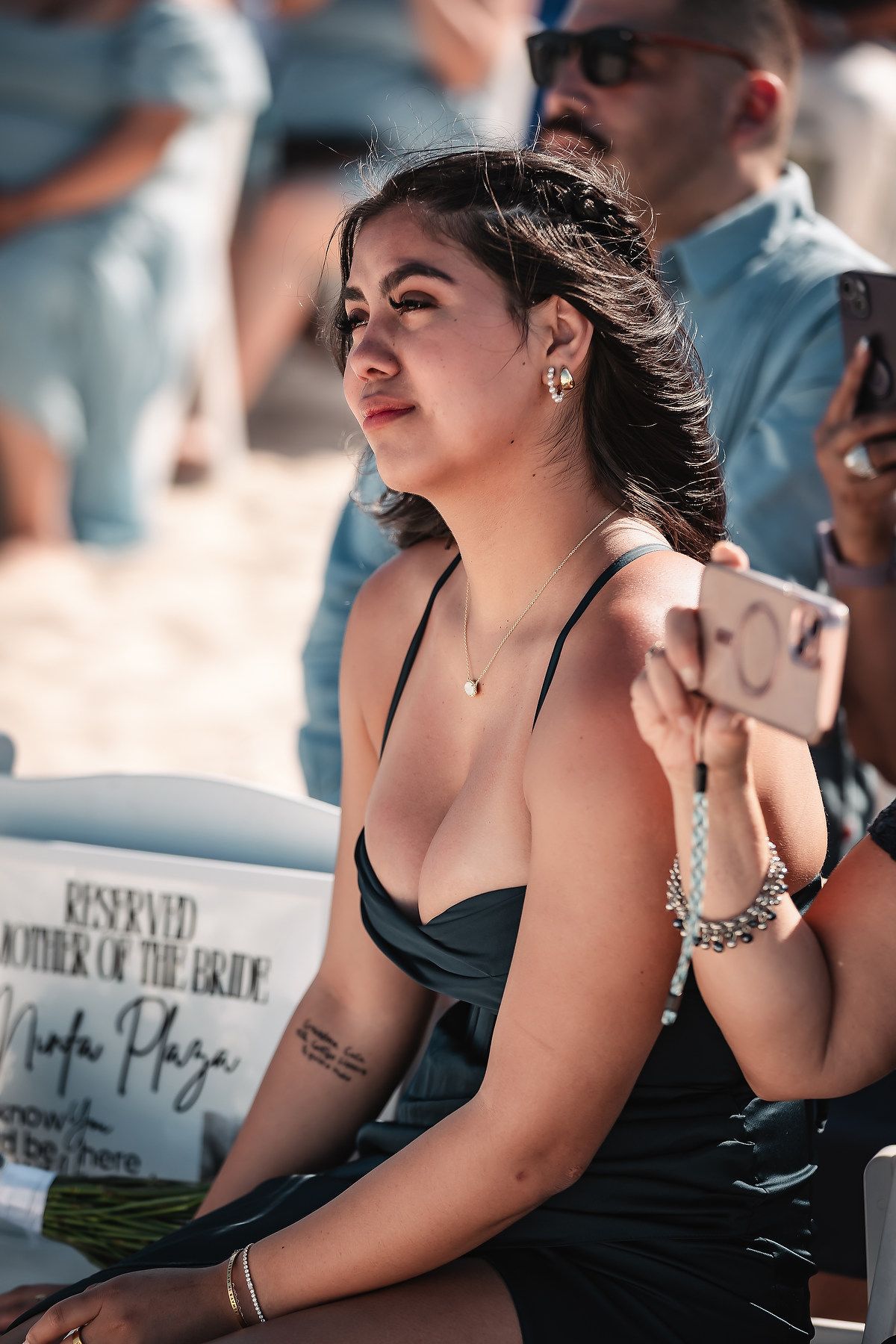 Woman in black dress sits at outdoor wedding, holding phone, with reserved Mother of the Bride sign nearby.