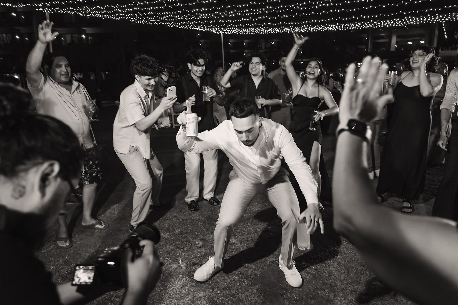 Black and white photo of a man dancing energetically at a wedding reception surrounded by cheering guests under string lights.