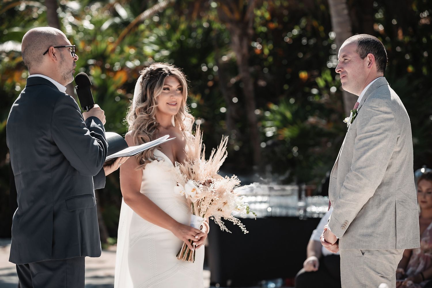 Bride holding dried floral bouquet exchanges vows with groom at outdoor wedding ceremony surrounded by lush greenery.