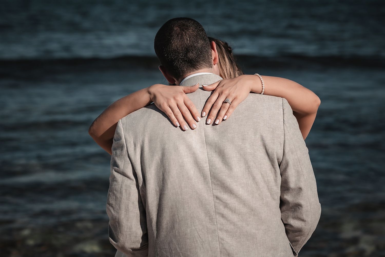 Couple embracing tenderly by the ocean shore, viewed from behind, wearing elegant beige outfits.