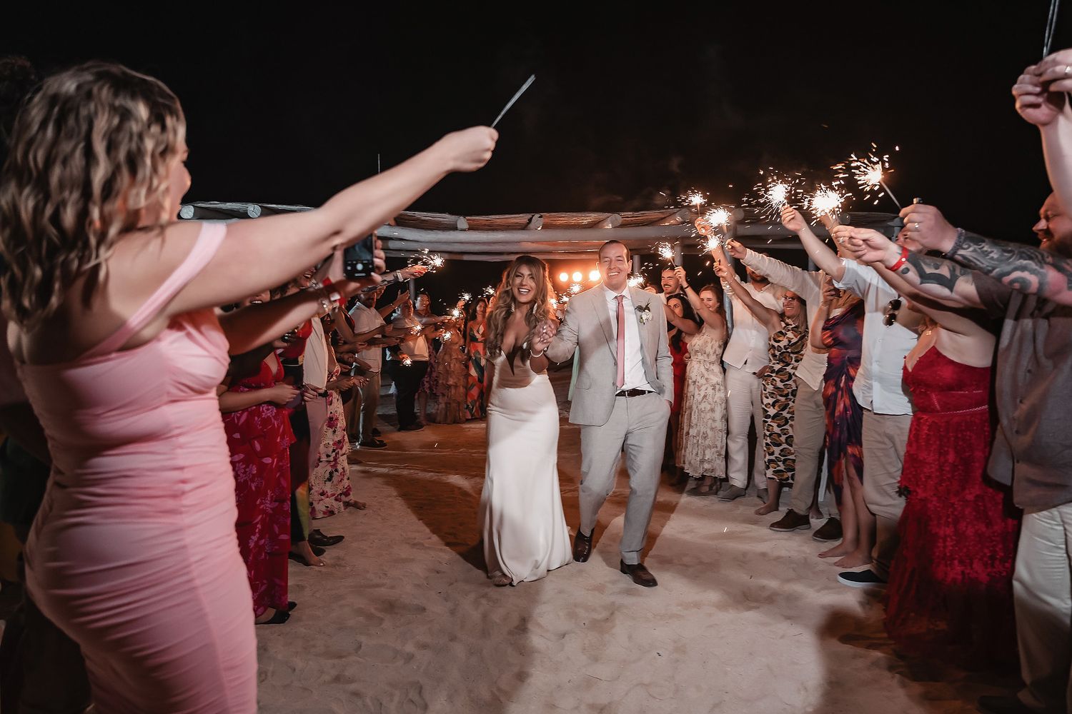 Bride and groom walk through sparkler exit surrounded by cheering wedding guests at night.