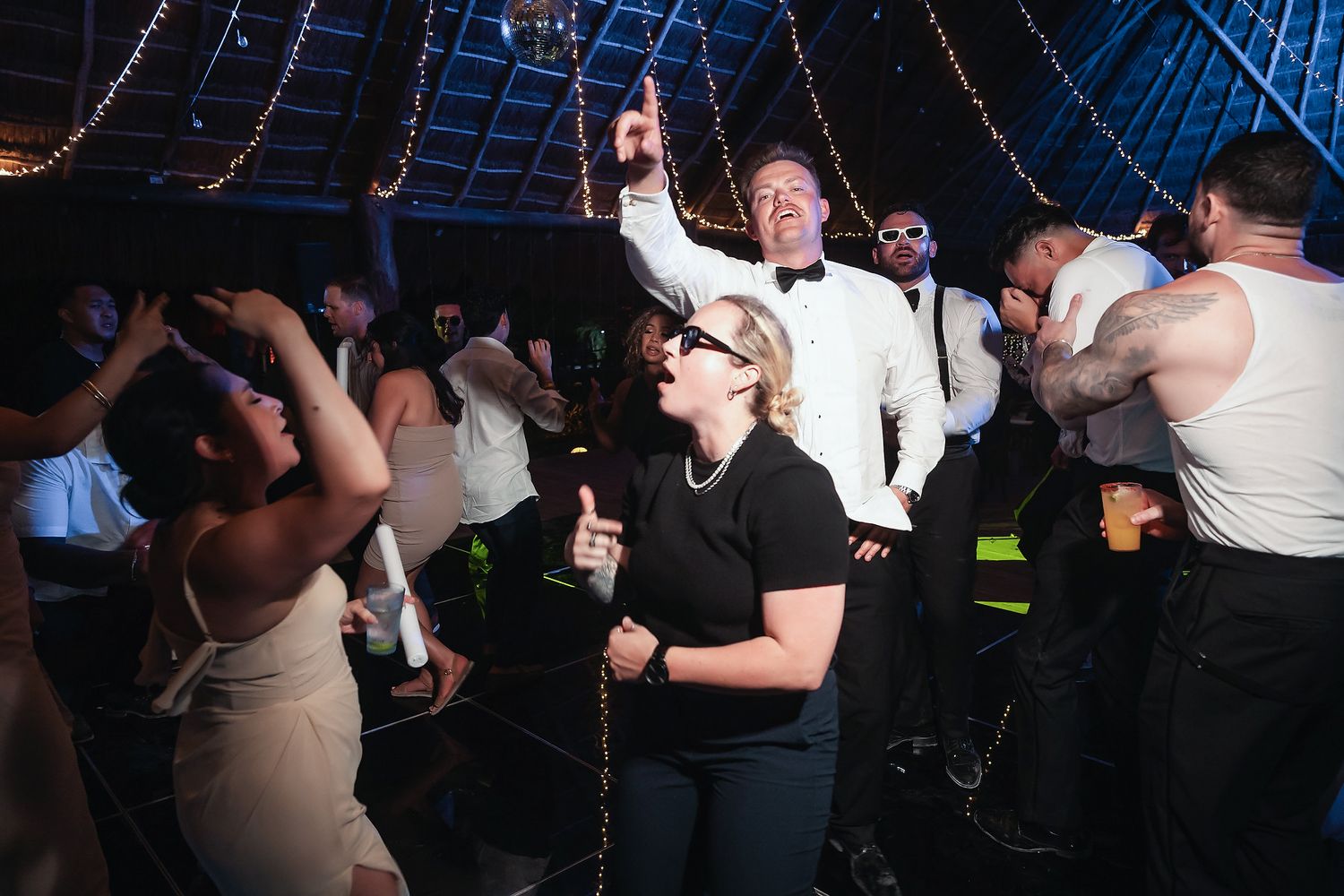 Guests dancing and celebrating joyfully at a lively wedding reception under twinkling string lights.