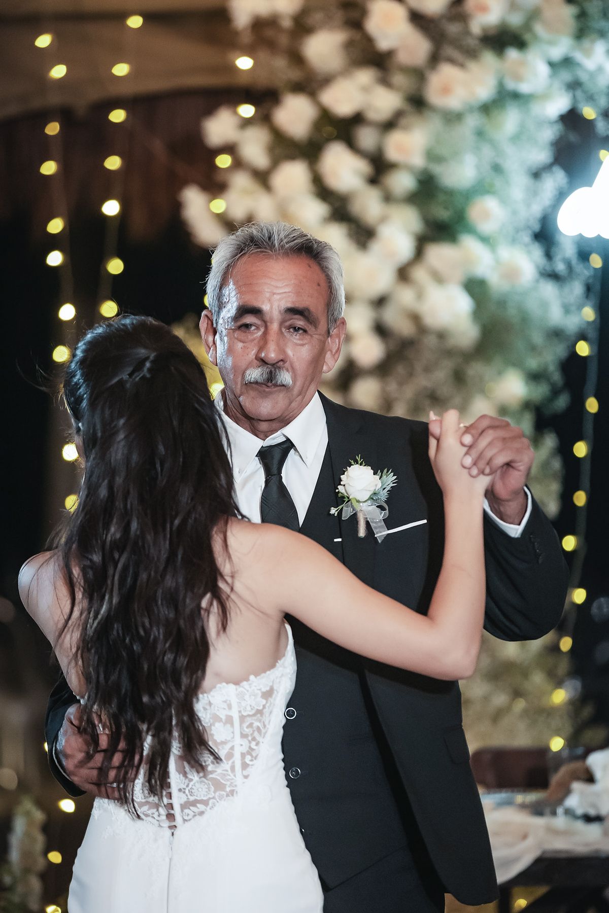 Bride and her father share an emotional first dance at a wedding reception with floral arrangements and string lights.