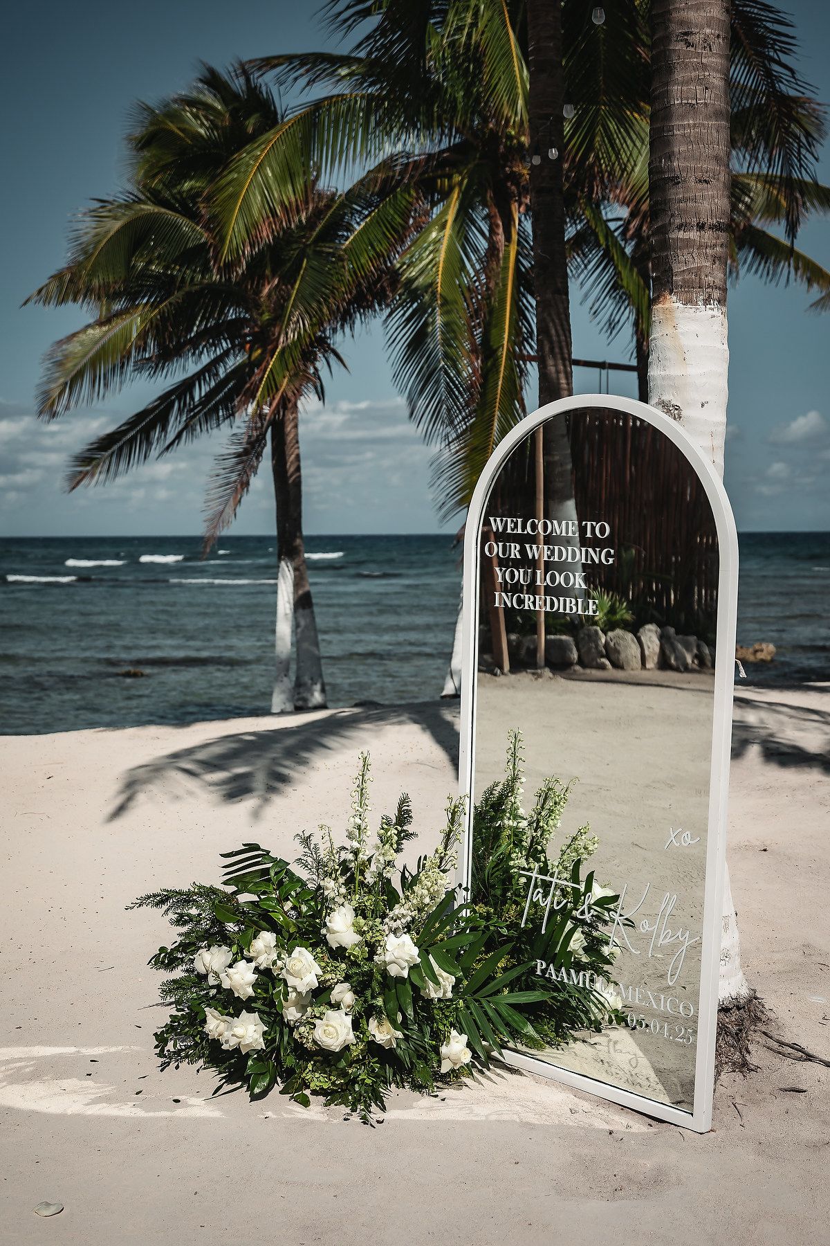 Elegant beach wedding welcome mirror with white floral arrangement, palm trees, and ocean backdrop on sandy shore.