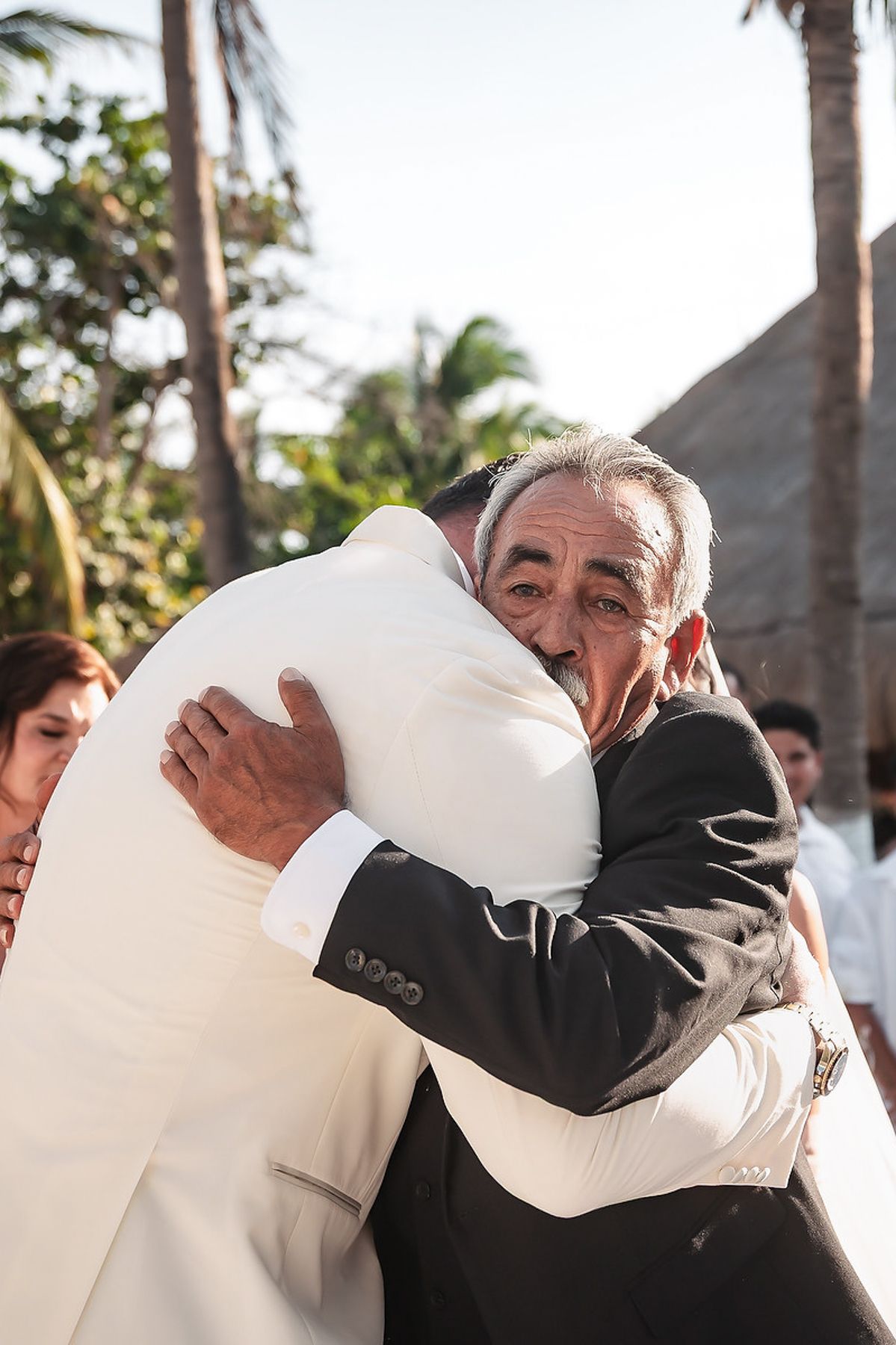 Emotional father embraces wedding guest in heartfelt hug during outdoor beach wedding celebration.