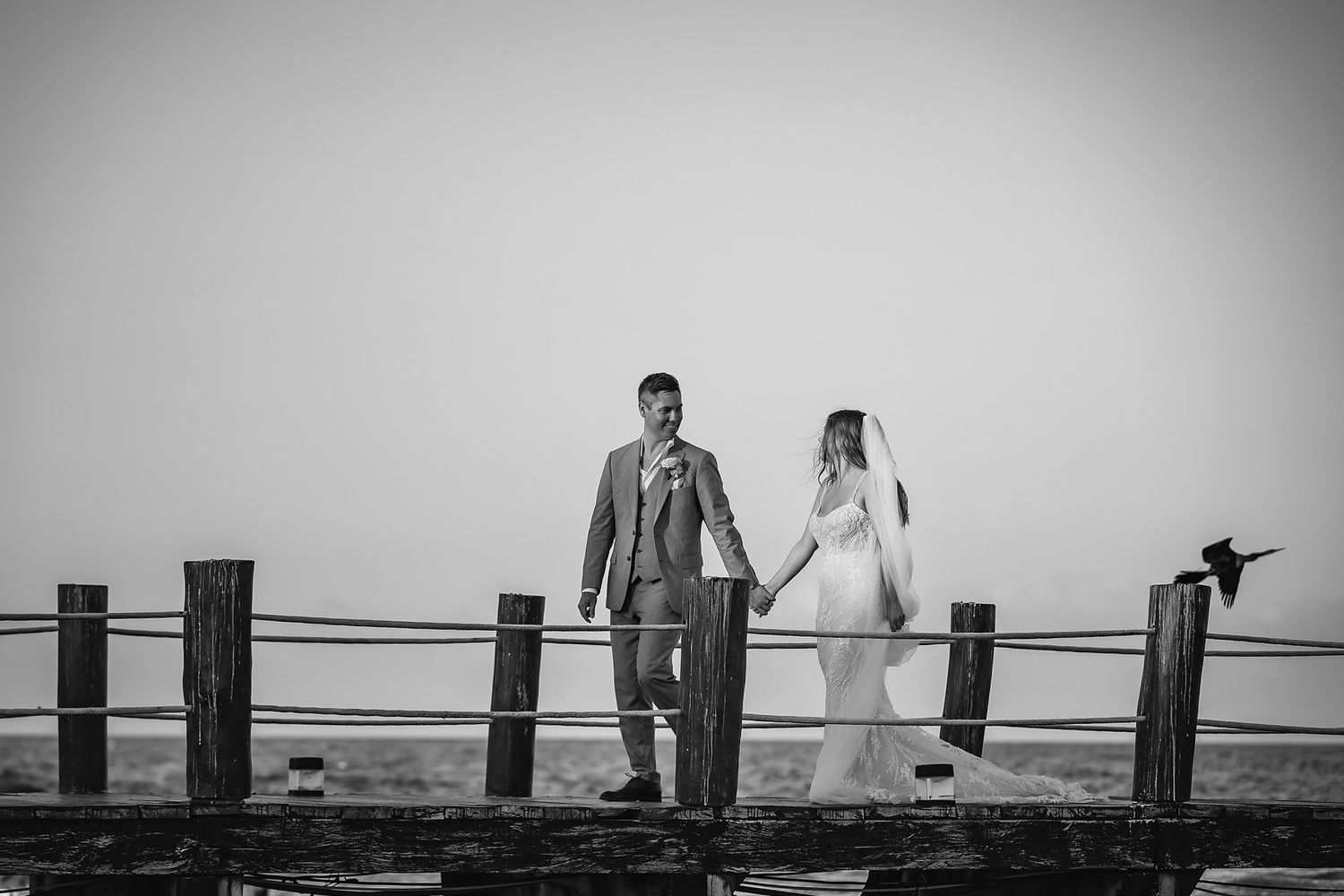 Black and white photo of bride and groom holding hands on a wooden pier fence at their beach wedding.