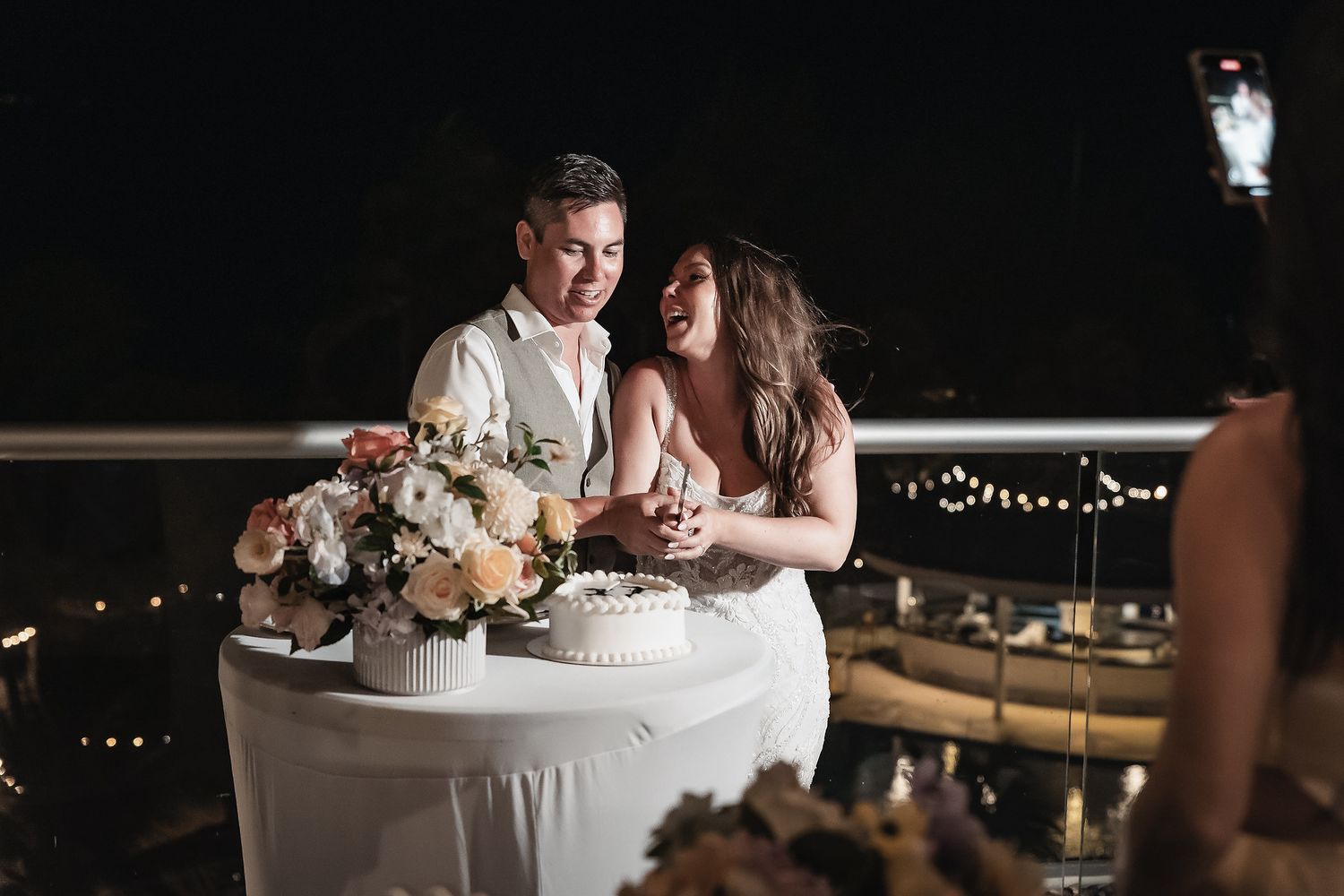Couple cutting wedding cake at night outdoor reception with floral arrangements and string lights in background.