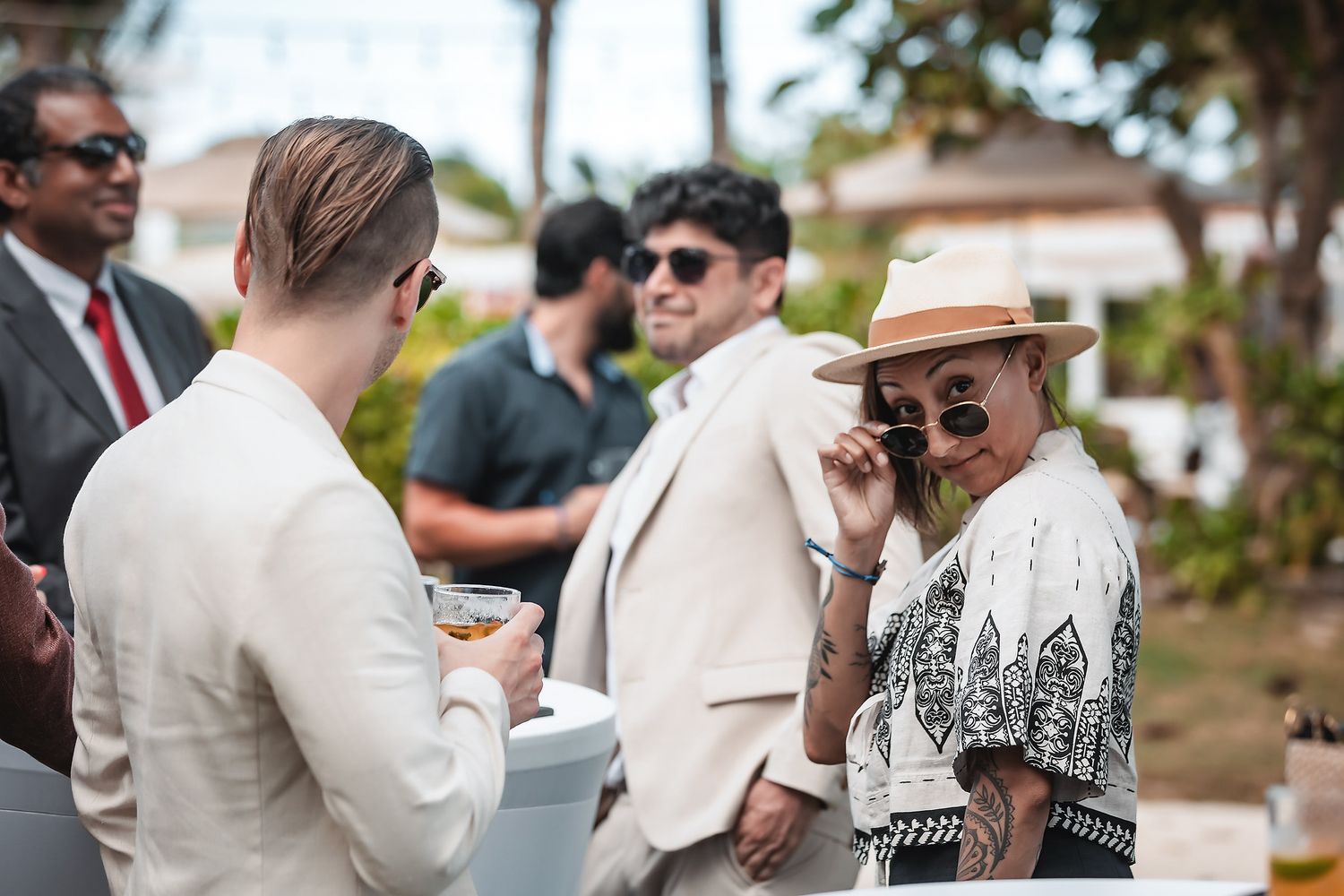 Elegantly dressed guests socializing outdoors at a tropical event, woman in hat and sunglasses smiling at man in beige suit.