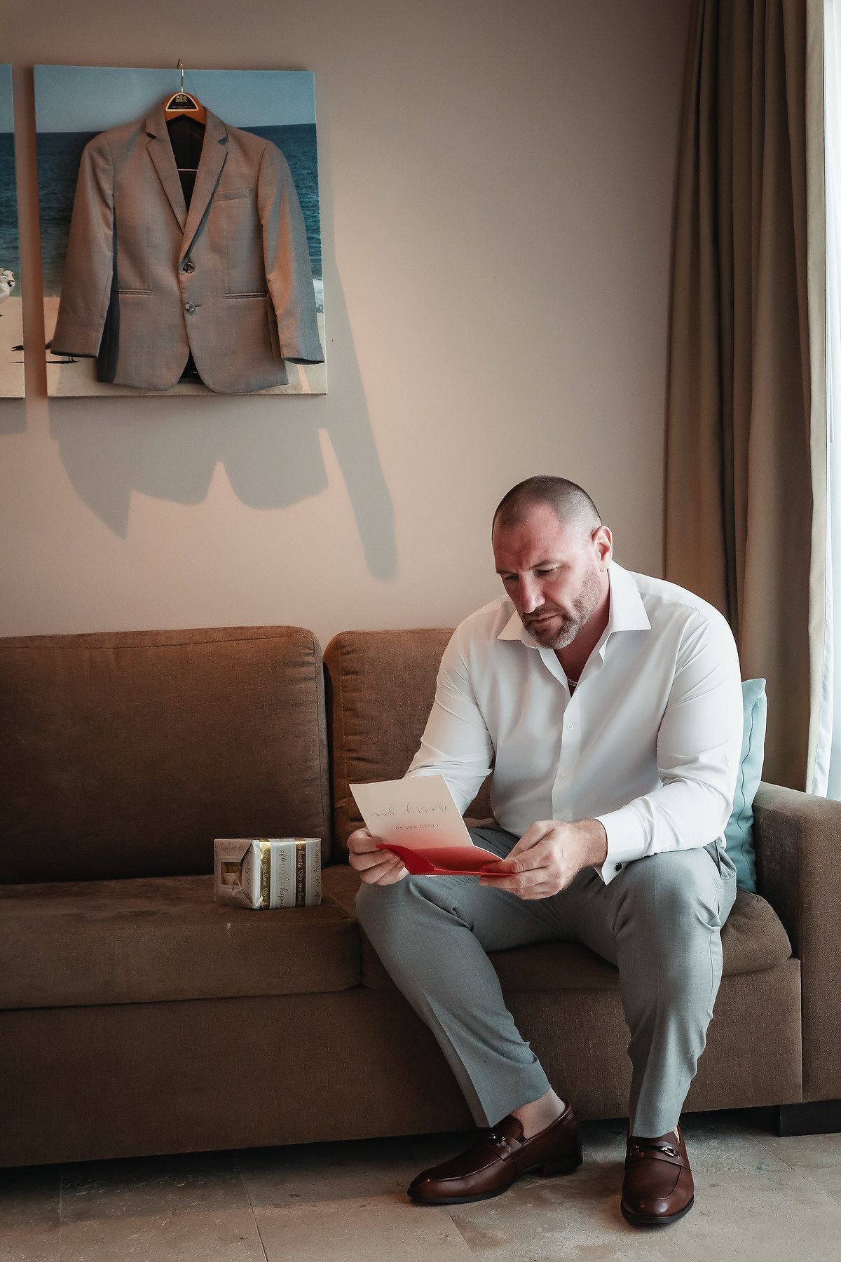Man in white shirt reading a red letter on a brown sofa with a suit jacket hanging on the wall.