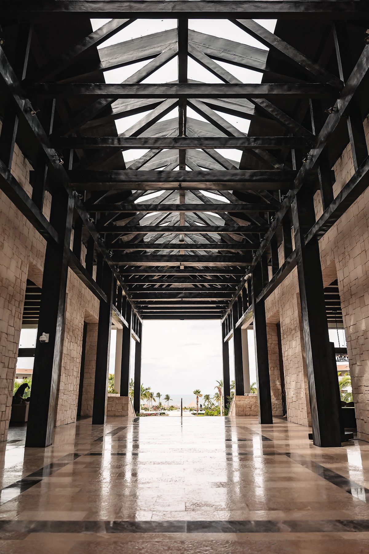 Grand open-air corridor with dark wooden beam ceiling, marble floors, and a scenic ocean view at a luxury resort.