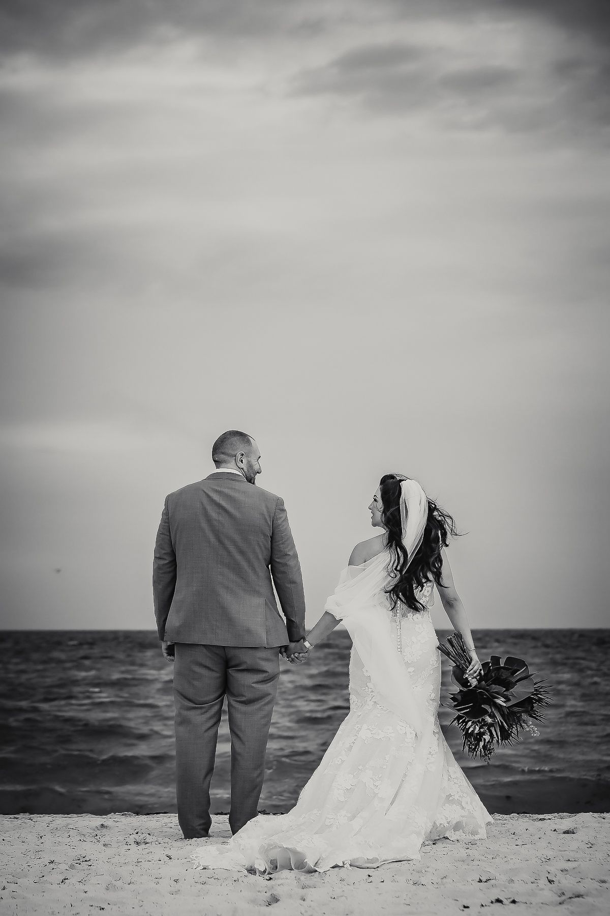 Black and white photo of bride and groom holding hands on beach, bride holding bouquet, ocean in background.