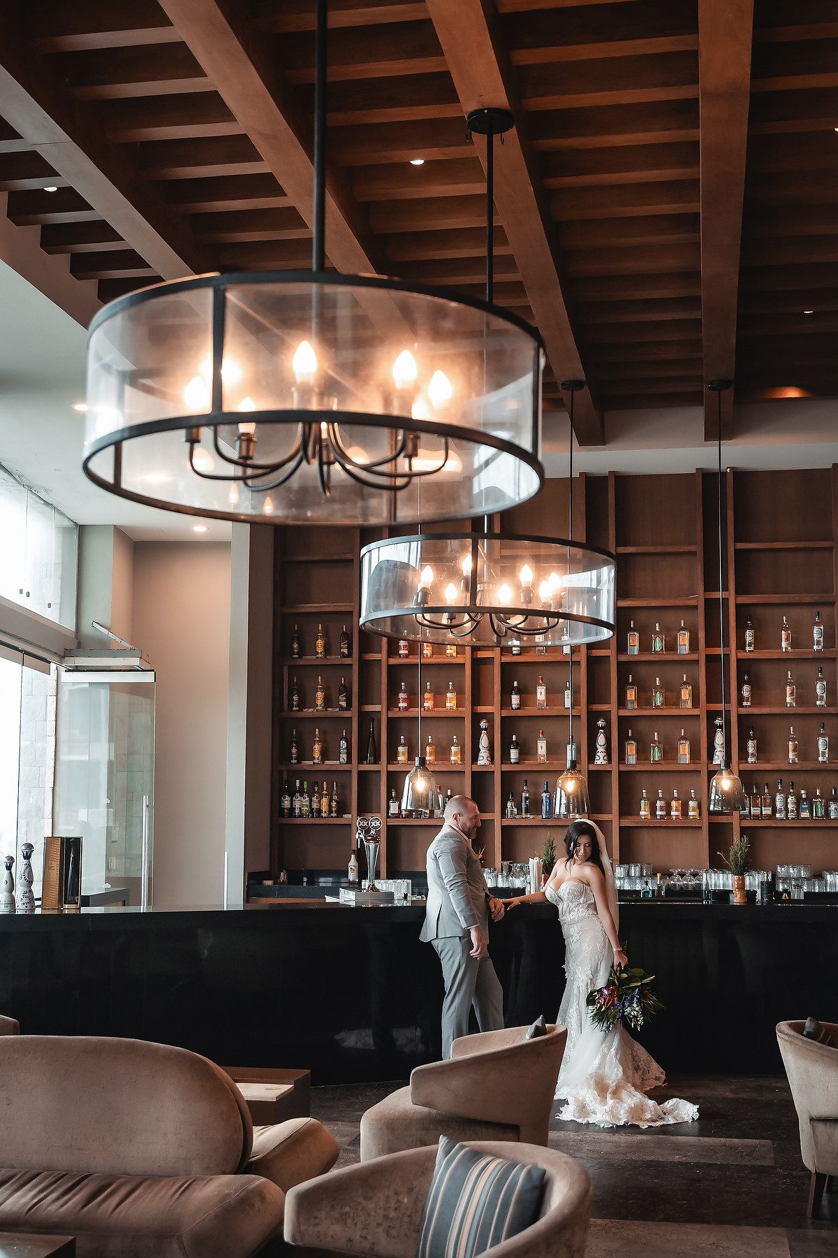 Bride and groom hold hands in an elegant hotel bar with wood shelving, pendant chandeliers, and warm ambient lighting.