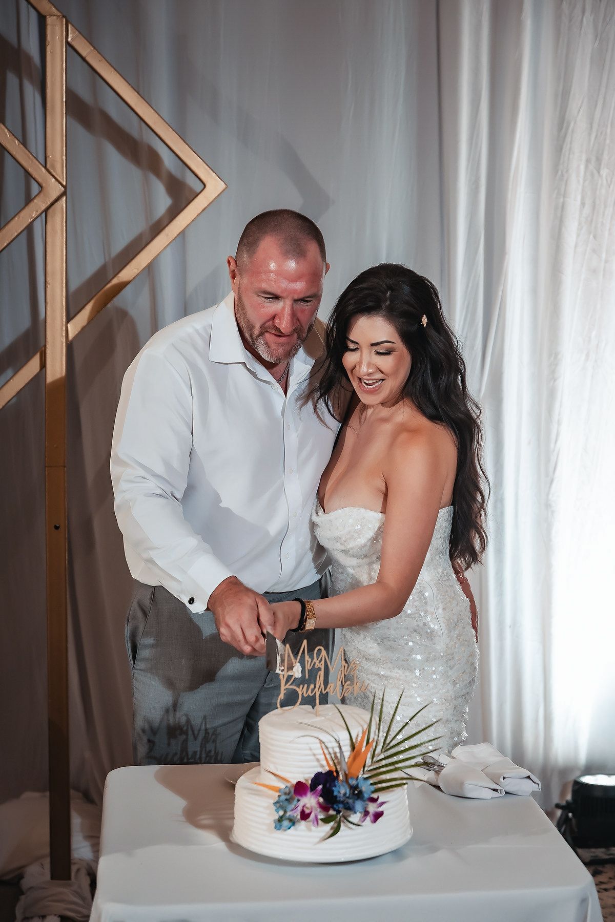 Smiling bride and groom cut their tropical-themed white wedding cake together during their reception celebration.