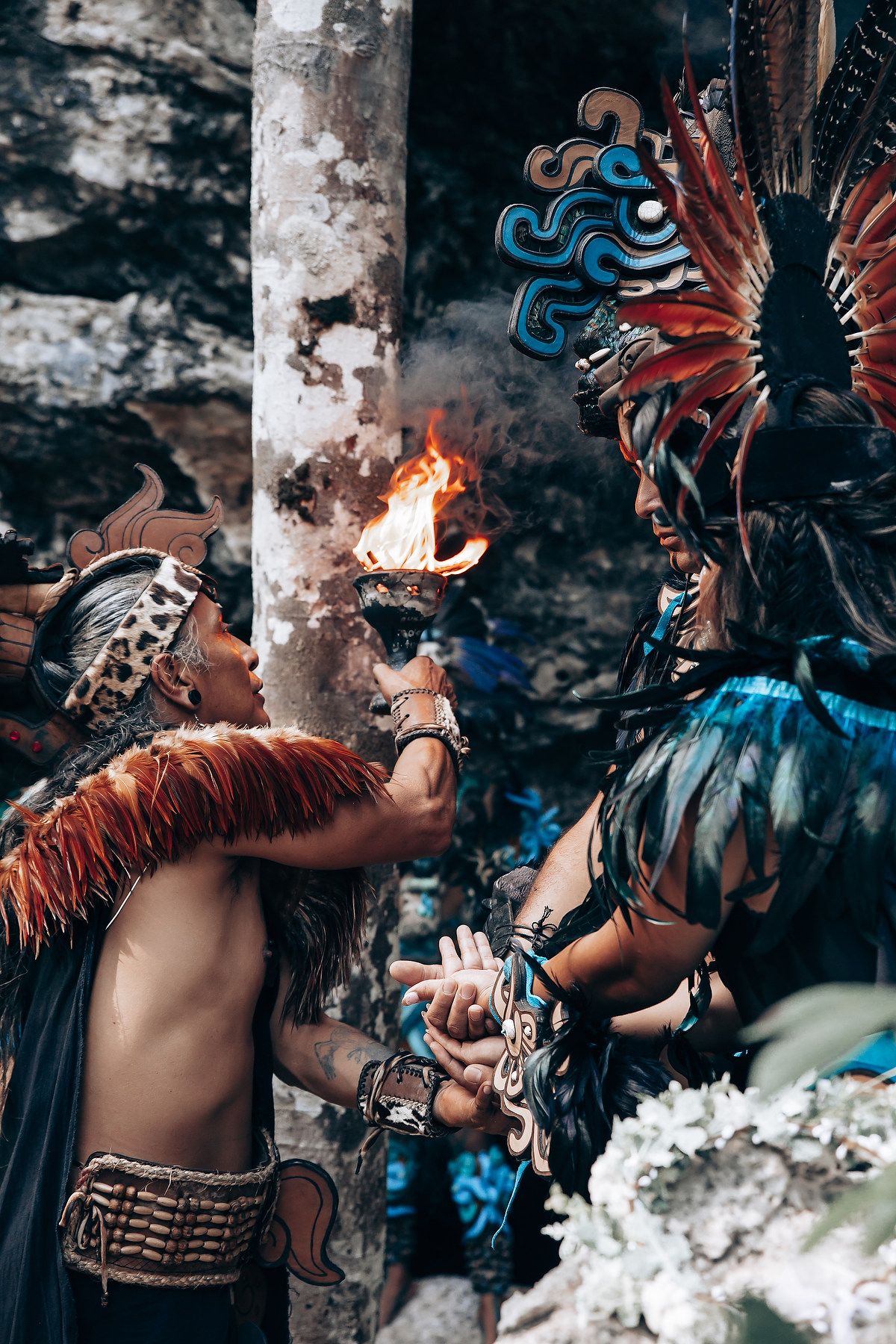 A shirtless man in Mayan attire holds a flaming torch toward a person in an elaborate feathered headdress.