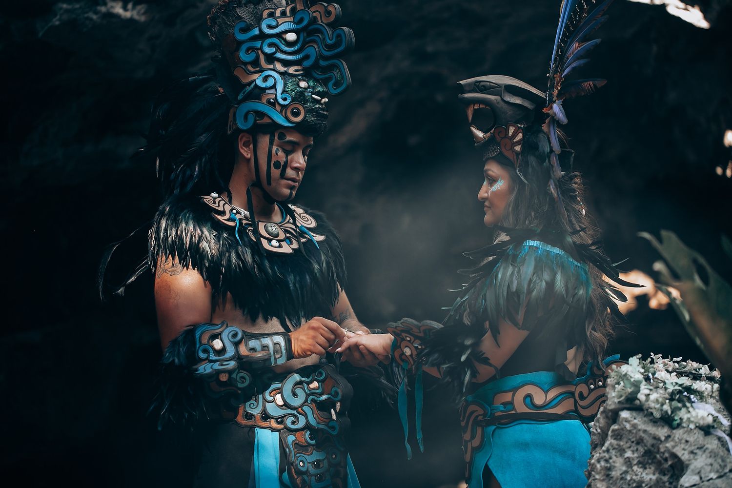 Two performers in blue Mayan ceremonial attire with feathered headdresses interacting in a misty cave setting.