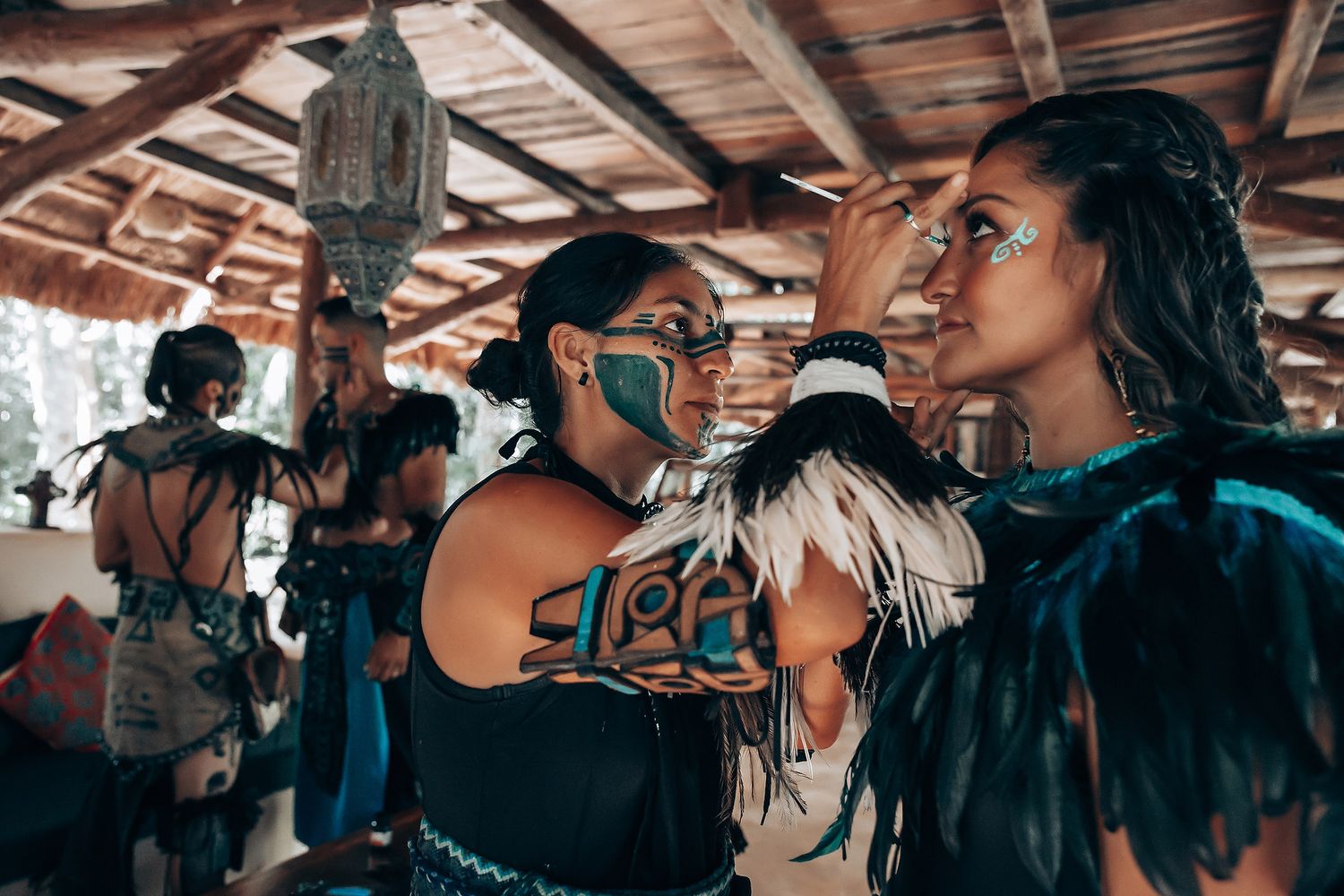 Women in traditional Mayan attire applying face paint in teal and white during cultural ceremony preparation.