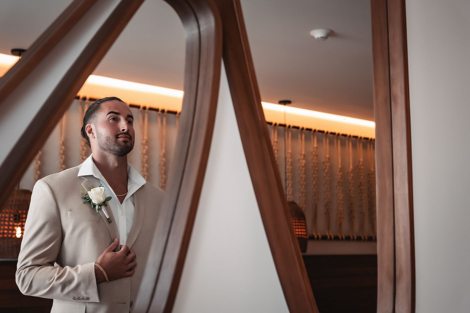 Elegant groom in beige suit with white boutonniere adjusting jacket near decorative wooden mirror frame.