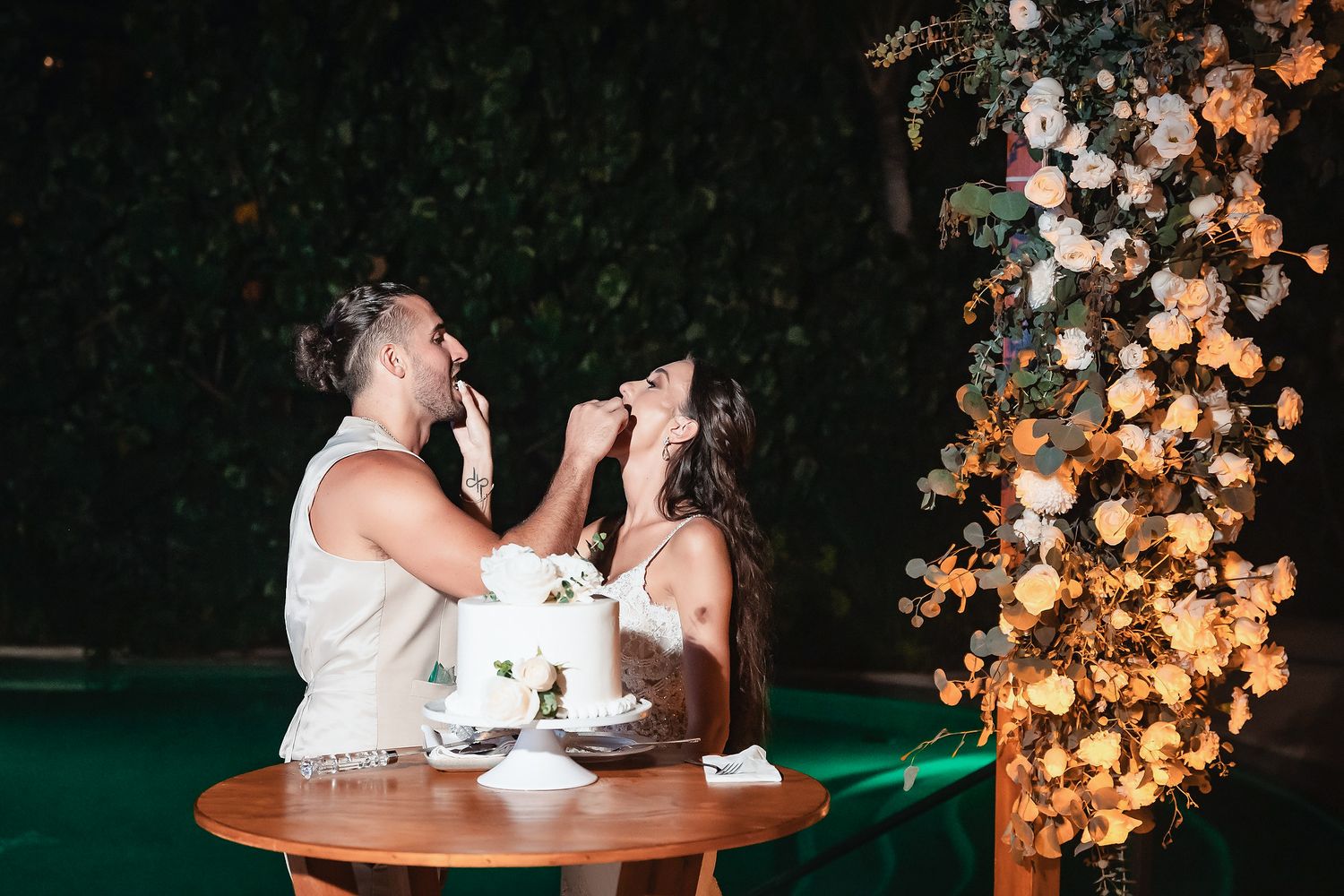 Bride and groom feeding each other wedding cake at night beside a floral arrangement of peach roses.