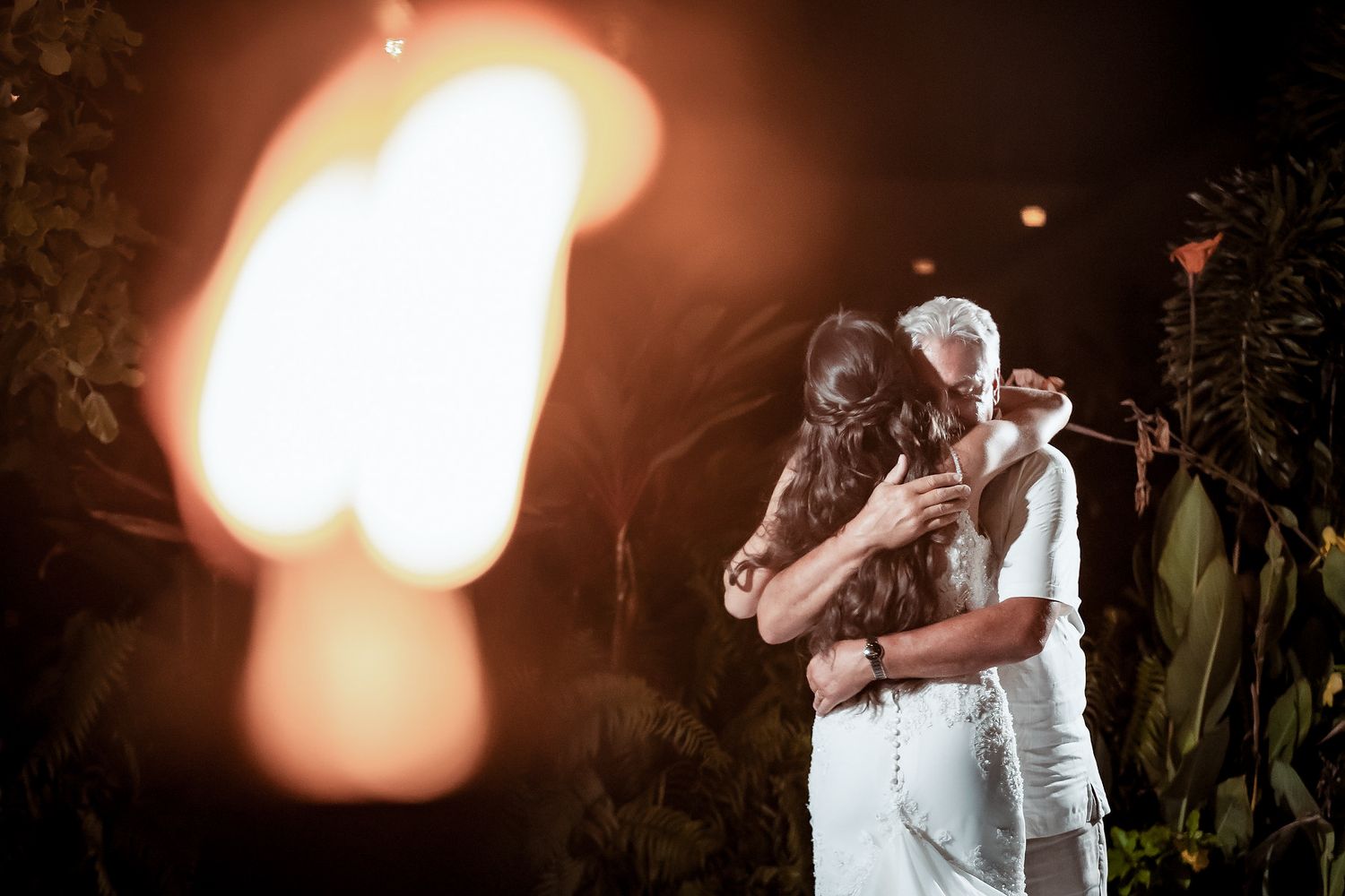 Couple sharing romantic embrace at night wedding ceremony with warm glowing lantern light and tropical greenery backdrop.