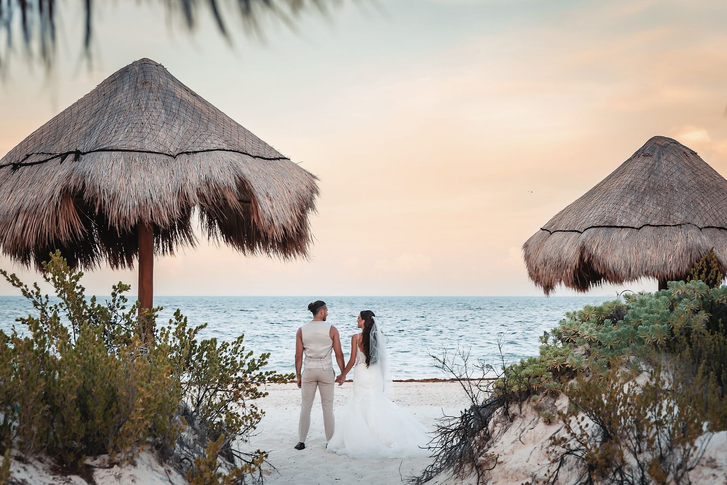 Couple holding hands on a sandy beach at sunset, framed by two thatched palapa umbrellas overlooking the ocean.