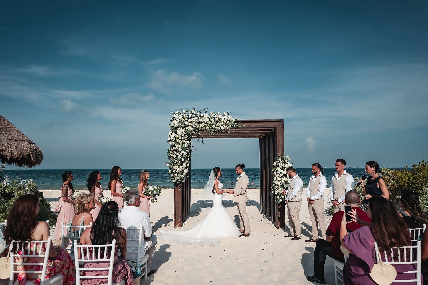 Beach wedding ceremony with floral arch, bride and groom, bridal party, and guests seated on white sand with ocean backdrop.